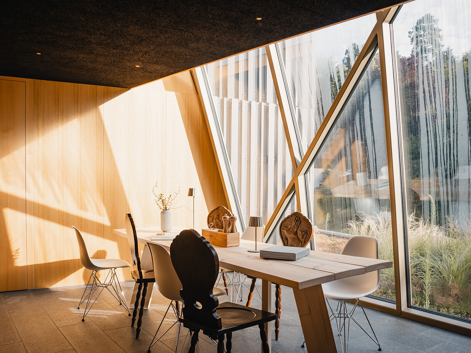Ein modern eingerichteter Raum mit großen, schrägen Fenstern, die natürliches Licht hereinlassen. Es gibt einen Tisch mit mehreren Stühlen, und die Wände sind aus hellem Holz. Dekorative Pflanzen sind vorhanden, und der Ausblick zeigt Bäume im Freien.