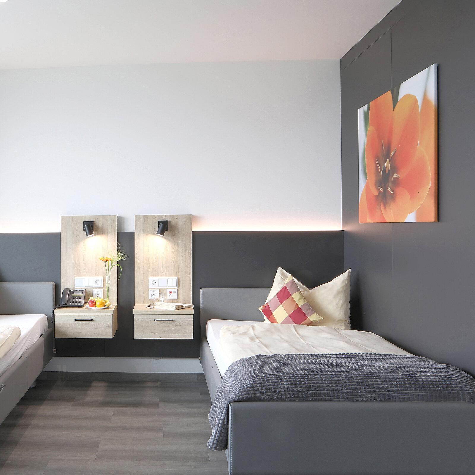 Das Bild zeigt ein modernes, helles Hotelzimmer mit zwei Einzelbetten, einer grauen Wand und einem großen Fenster. An der Wand sind zwei Nachtkästchen mit Lampen und einer Blume dekoriert. Ein großes Bild einer Blume hängt an der rechten Wand.