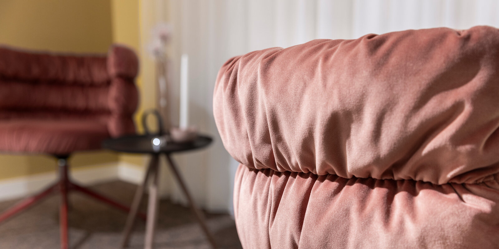 Close-up of a plush, pink cushioned chair with a blurred background featuring another chair, a small round table, and a vase.
