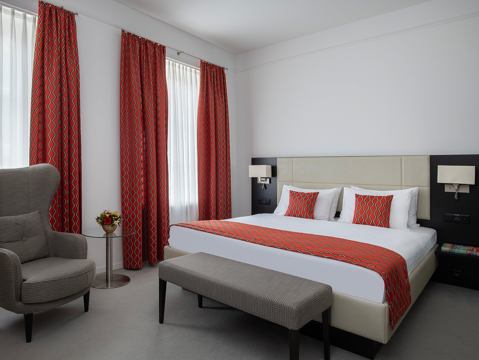 Ein modernes Hotelzimmer mit einem großen Doppelbett, das weiße Bettwäsche und rote, gemusterte Akzente hat. Zwei Fenster mit roten Vorhängen, ein beiger Sessel, ein brauner Hocker am Bettende und ein kleiner Tisch mit Blumen ergänzen die Einrichtung.