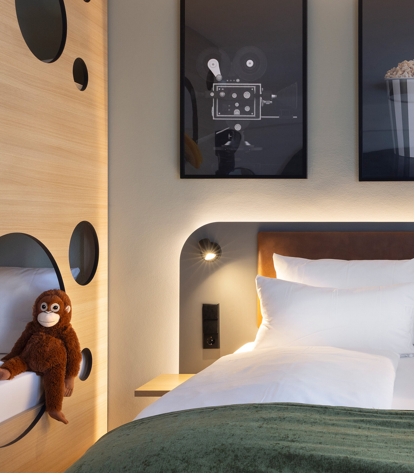 A cozy hotel room featuring a neatly made bed with white linens and a green throw. On the bedside is a stuffed monkey, and the wall displays two framed art pieces. The ambiance is modern and welcoming, with circular cutouts and soft lighting.