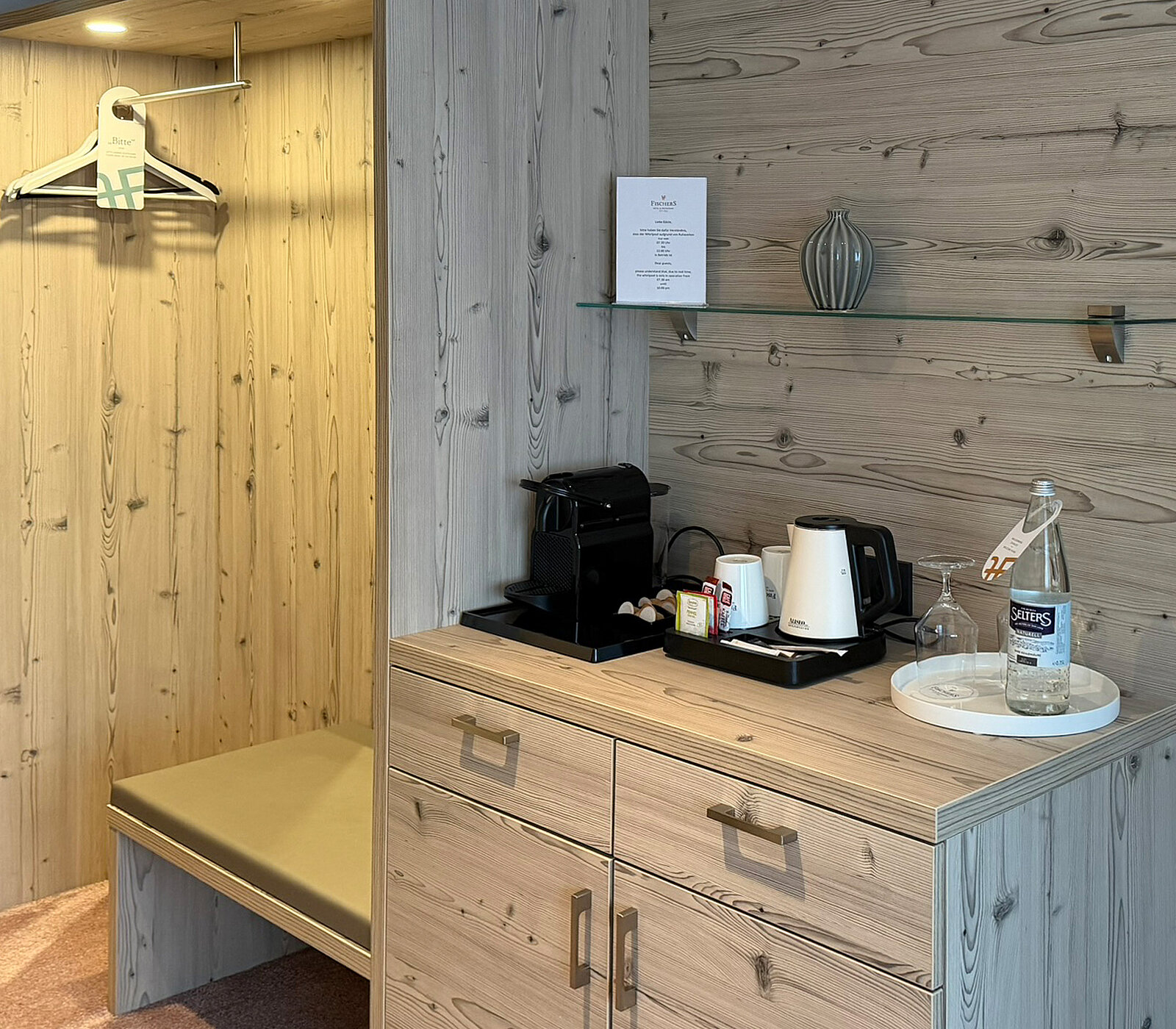 The image shows a wooden closet with a hanger, and next to it, a countertop featuring a coffee machine, kettle, and cups. A bottle of water and a small plant are displayed, alongside a menu on the wall. The overall setting is warm and inviting.