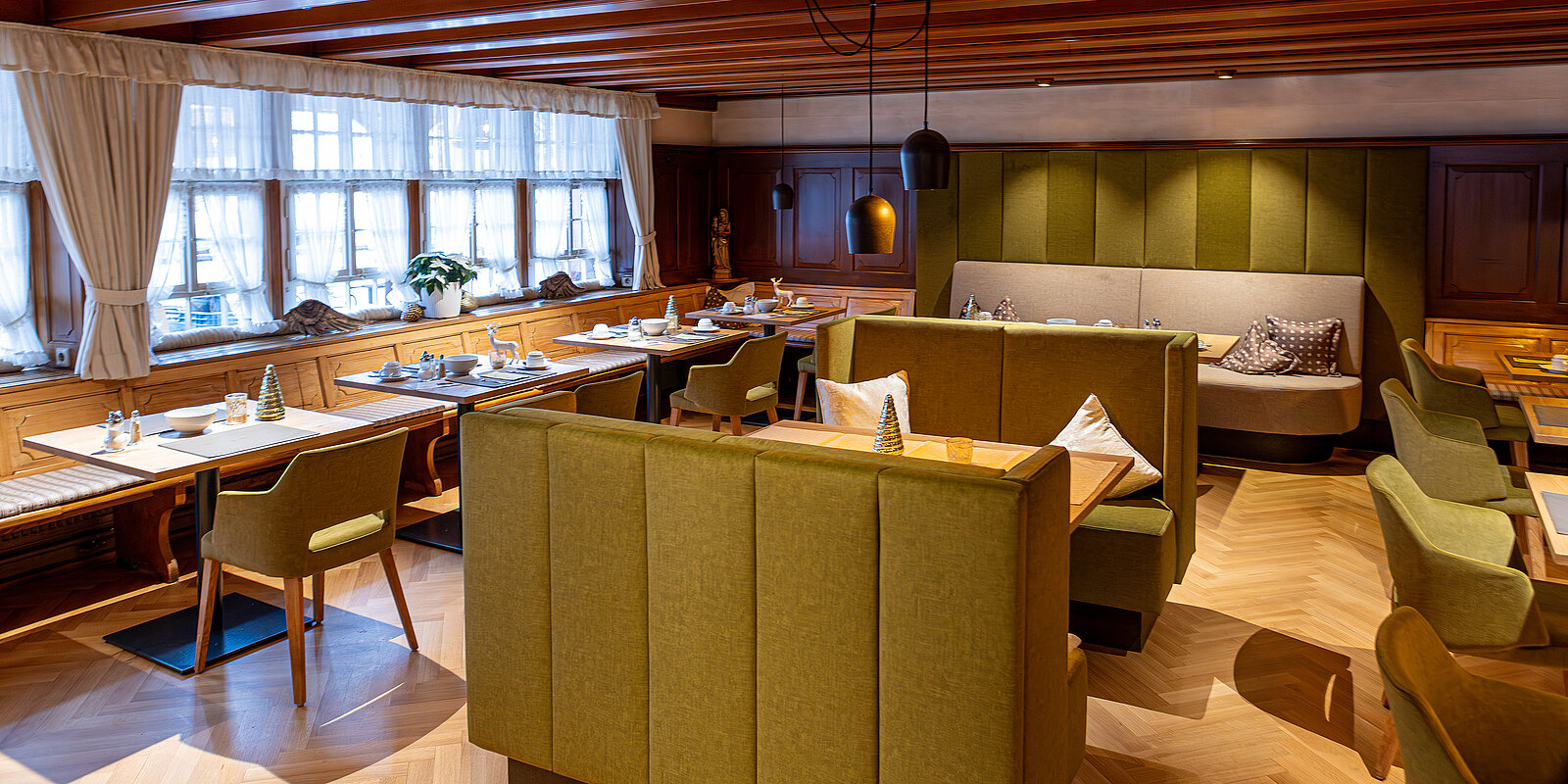 Gemütliches Restaurantinterieur mit Holzboden und grünen Polstermöbeln. Mehrere gedeckte Tische mit Stühlen und Bänken. Große Fenster mit Vorhängen lassen natürliches Licht herein. Elegante, ruhige Atmosphäre für ein entspanntes Essenserlebnis.