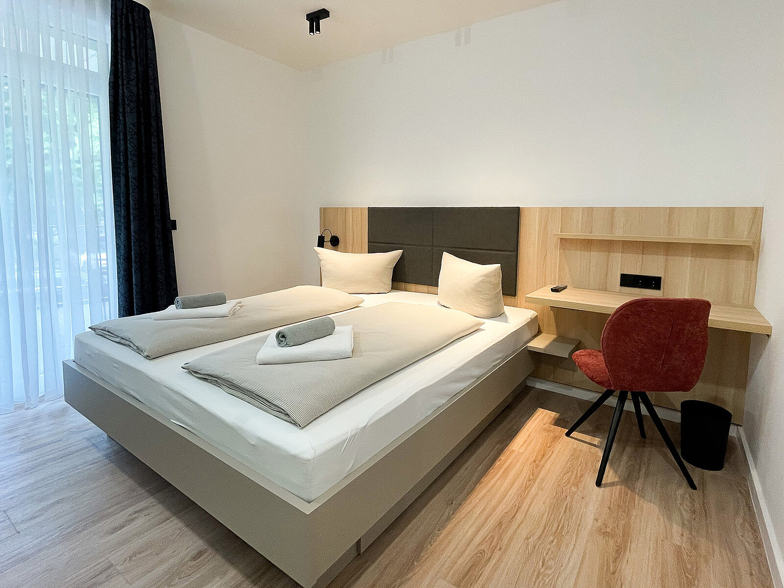 Ein modernes Schlafzimmer mit einem Doppelbett, das mit weißen Kissen und Decken ausgestattet ist. Daneben steht ein roter Stuhl an einem hellen Holzschreibtisch. Der Raum hat helle Wände und einen Holzboden.