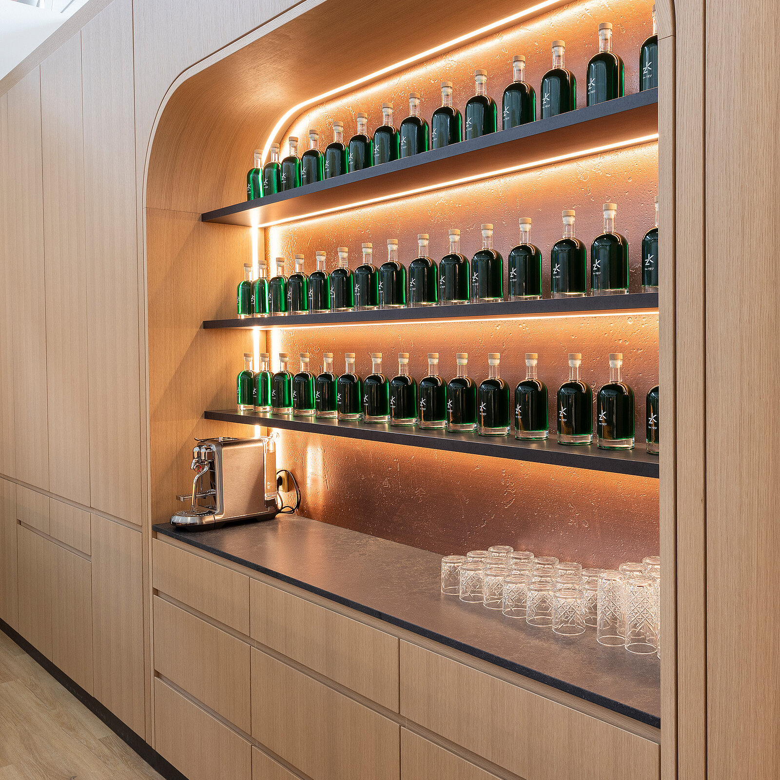 A modern display of green glass bottles arranged on wooden shelves, illuminated by soft lighting. Below the shelves, a wooden countertop features a beverage dispenser and clear glasses. The overall aesthetic is sleek and minimalist.