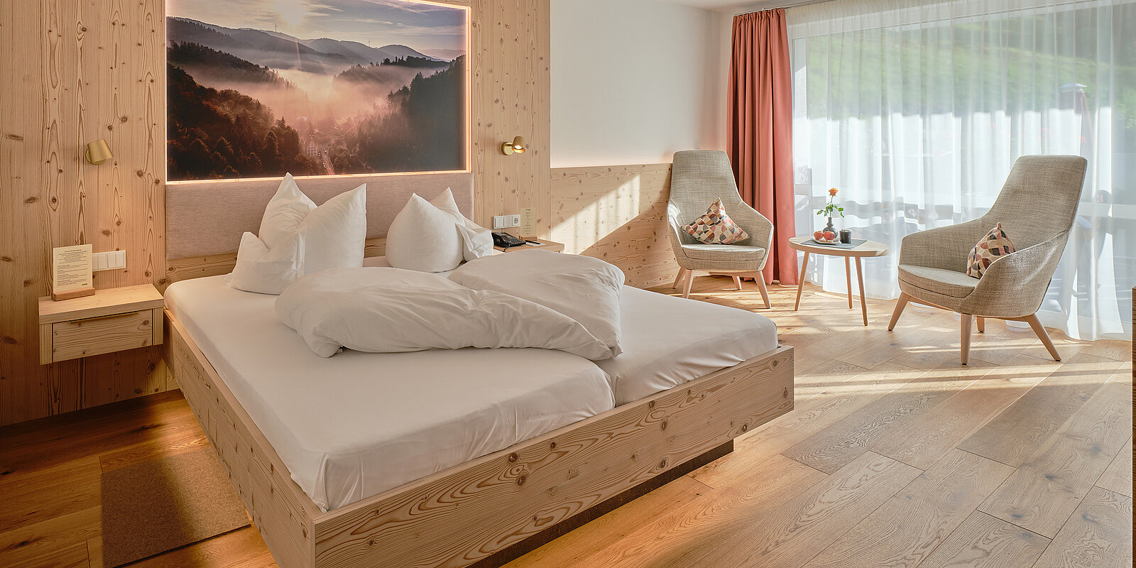 Gemütliches Hotelzimmer mit Holzmöbeln: ein großes Bett, zwei Sessel, ein kleiner Tisch mit Blume und Getränk. Warme Beleuchtung und ein Wandbild mit Berglandschaft. Große Fenster mit Vorhängen lassen viel Tageslicht herein.