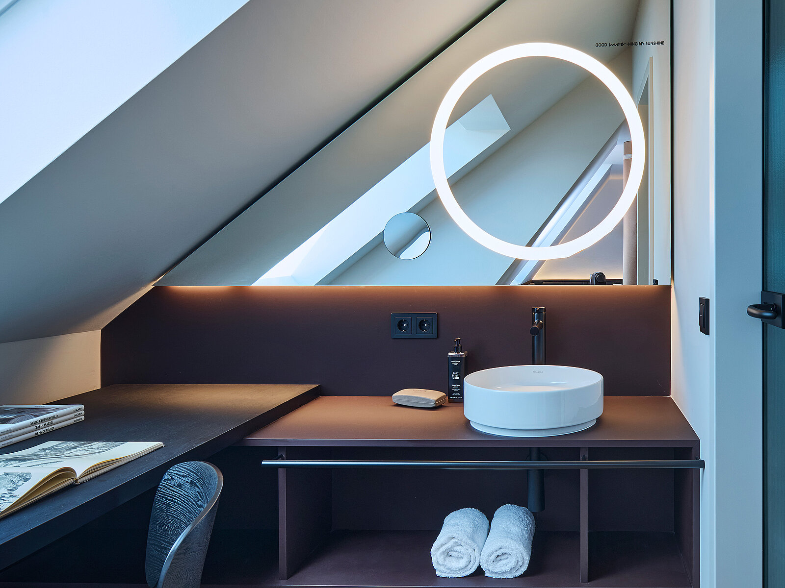 In einem modernen Dachzimmer befindet sich ein rundes Waschbecken mit einem beleuchteten Spiegel. Daneben sind Pflegeprodukte, ein kleiner Spiegel und gerollte Handtücher zu sehen. Das Ambiente ist minimalistisch und elegant gestaltet.
