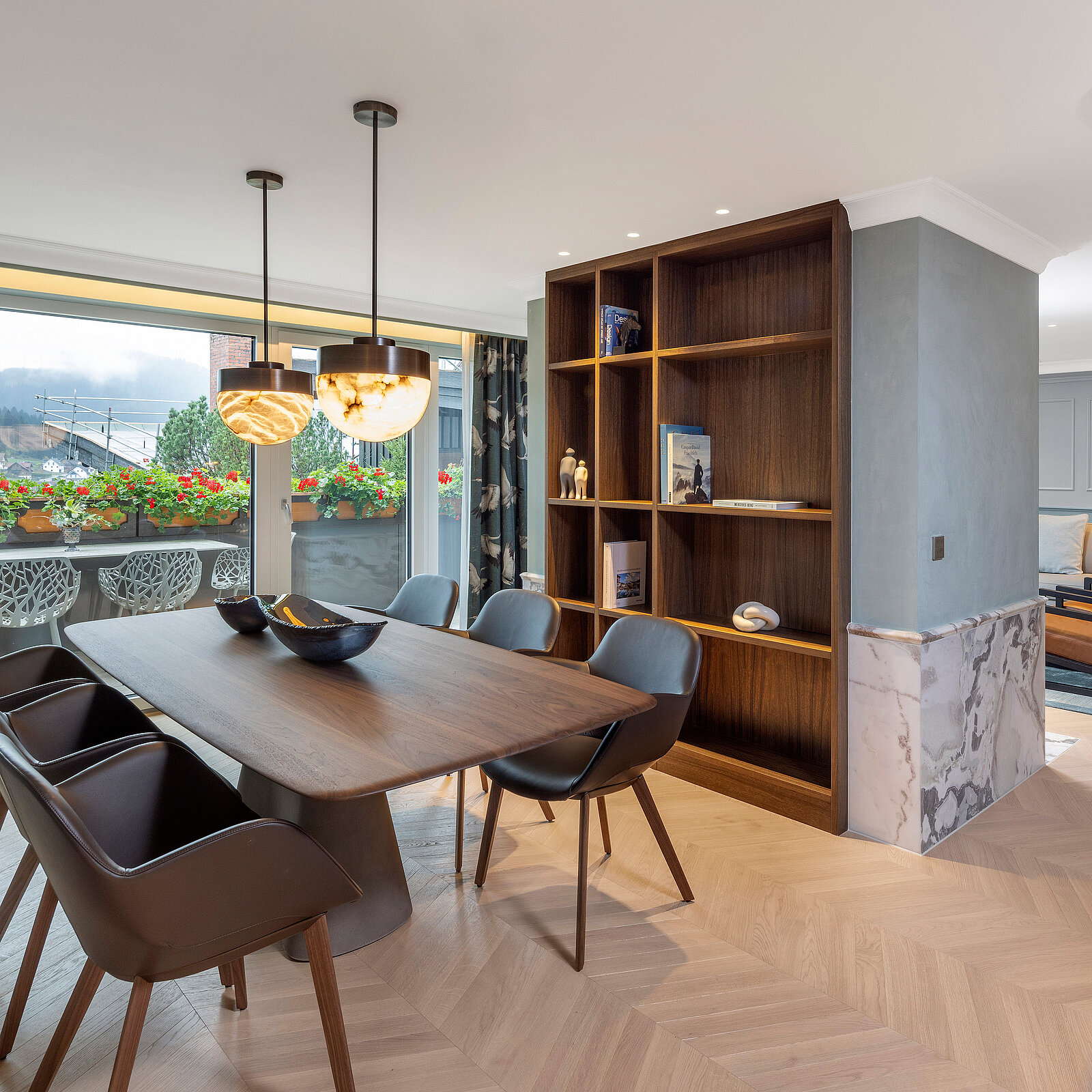 Ein moderner Essbereich mit einem großen Holztisch und eleganten Stühlen. Über dem Tisch hängen stilvolle Lampen. Im Hintergrund sind große Fenster, die natürliches Licht hereinlassen, sowie ein Bücherregal. Der Raum ist geschmackvoll dekoriert und vermittelt eine einladende Atmosphäre.