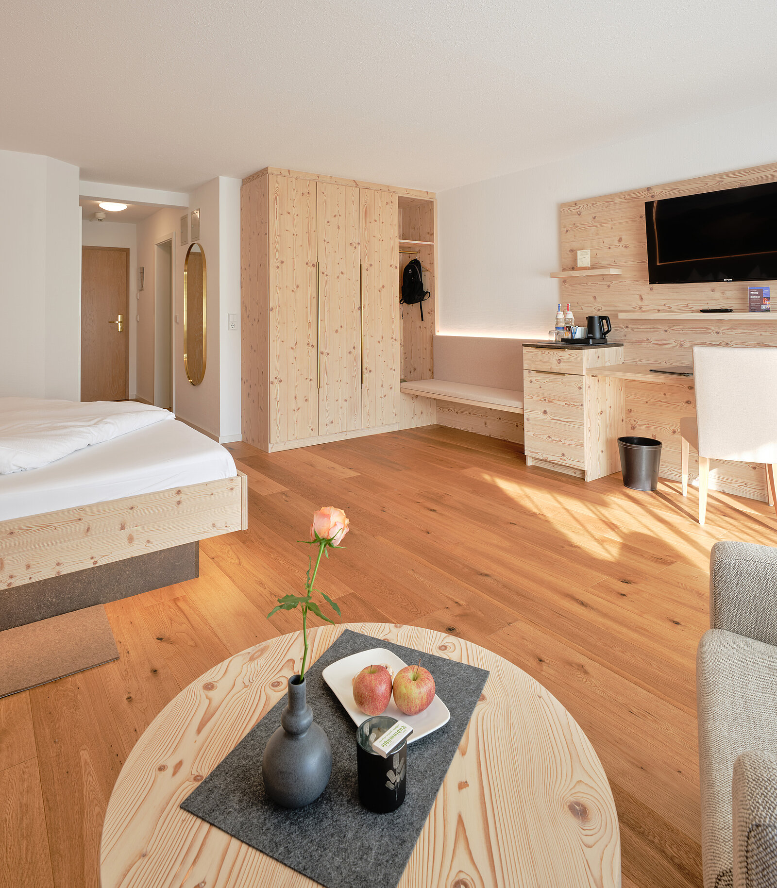 Ein modernes Hotelzimmer mit Holzmöbeln, einem Bett, einem Tisch mit Vase, zwei Äpfeln, einem Wasserkocher und einem Fernseher an der Wand. Der Raum wirkt hell und einladend durch das Holzdesign und die natürliche Beleuchtung.