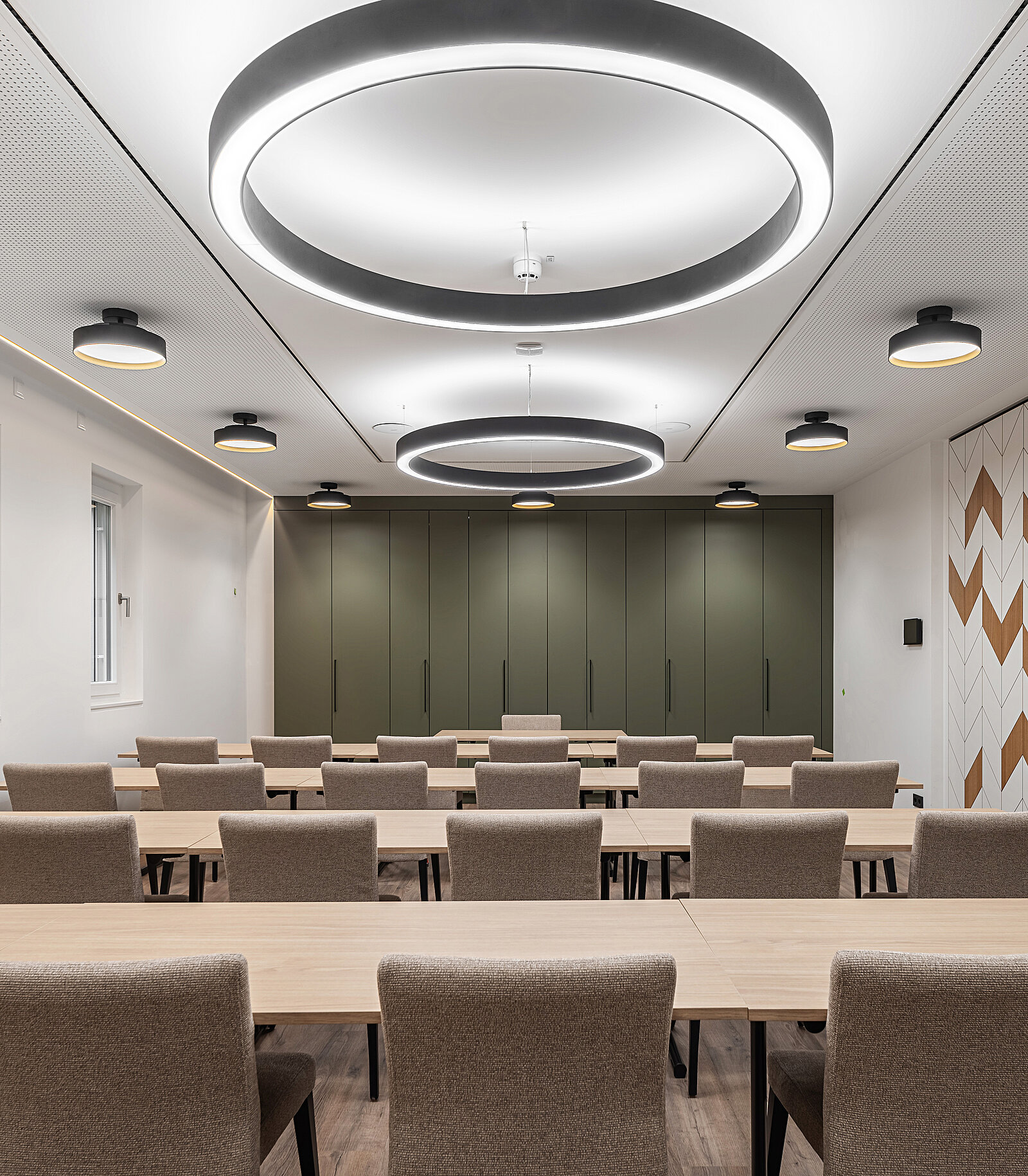 Ein moderner Tagungsraum mit mehreren Tischen und Stühlen. Die Wände sind in neutralen Farben gehalten, mit einem grünen Schrank im Hintergrund. Deckenleuchten mit runden und rechteckigen Formen sorgen für ausreichend Licht. Der Raum wirkt einladend und funktional für Meetings oder Schulungen.