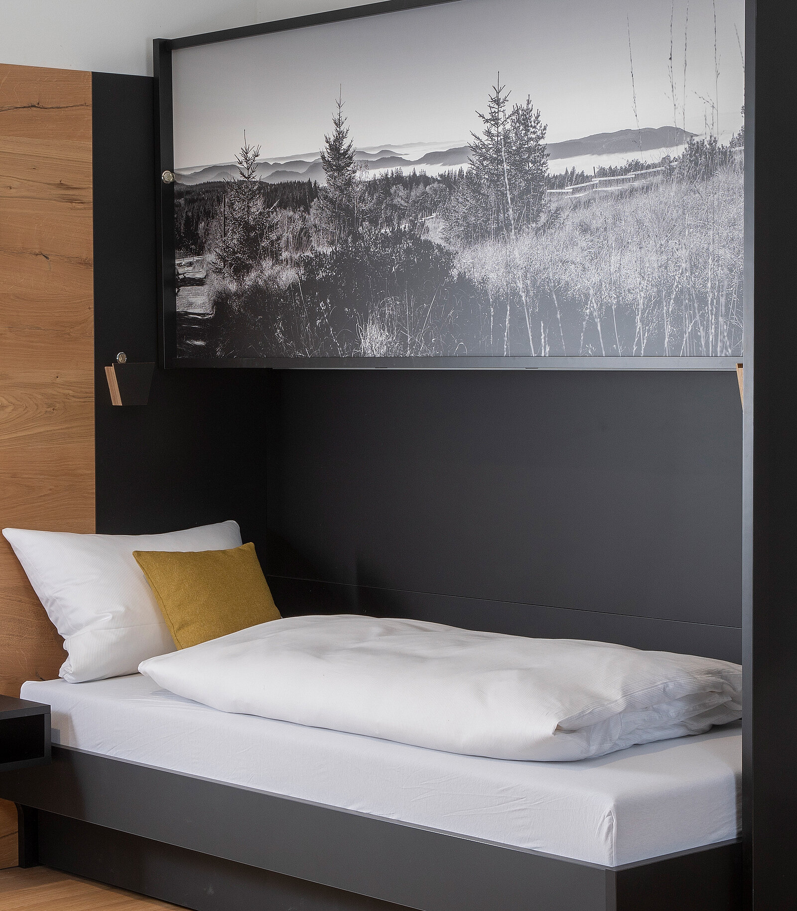 Ein einfaches Bett mit weißer Bettwäsche und einem gelben Kissen. Über dem Bett hängt ein großflächiges Schwarz-Weiß-Foto einer bewaldeten Landschaft mit Bergen im Hintergrund. Der Raum hat ein modernes Design mit Holz- und dunklen Wandelementen.