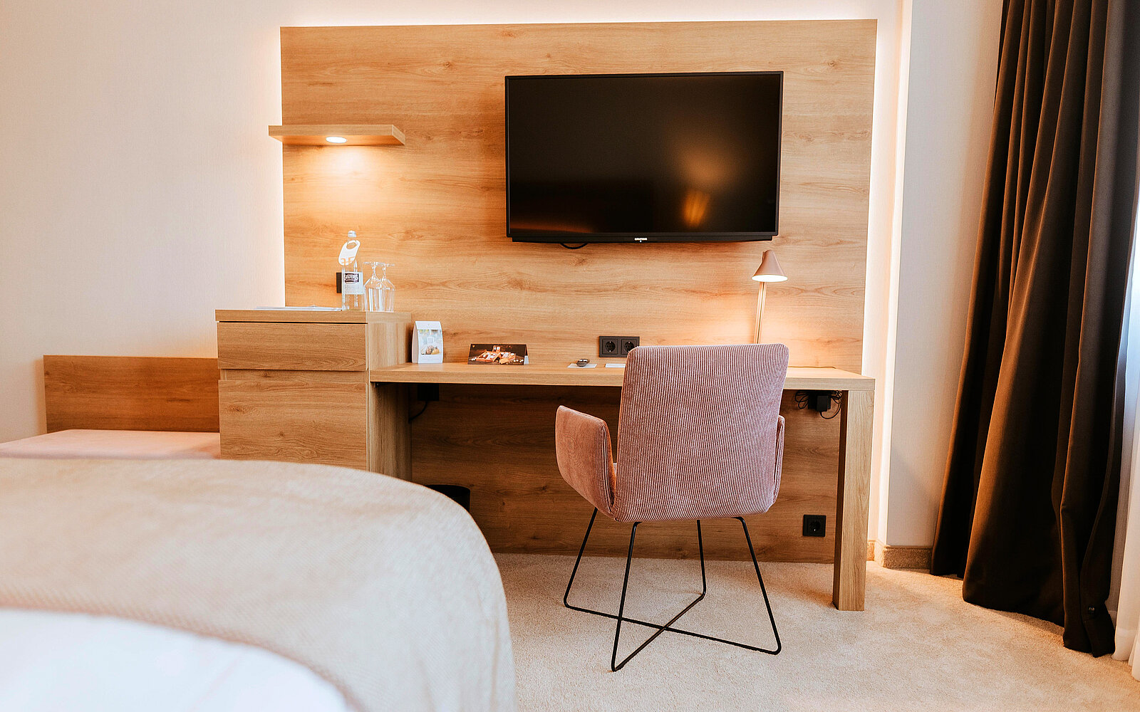 Das Bild zeigt ein modernes Hotelzimmer mit einem Holzpaneel, auf dem ein Fernseher montiert ist. Neben dem Fernseher steht eine kleine Lampe. Unter dem Fernseher befindet sich ein Schreibtisch mit einem pinkfarbenen Stuhl, einer Flasche Wasser und weiteren Gegenständen.
