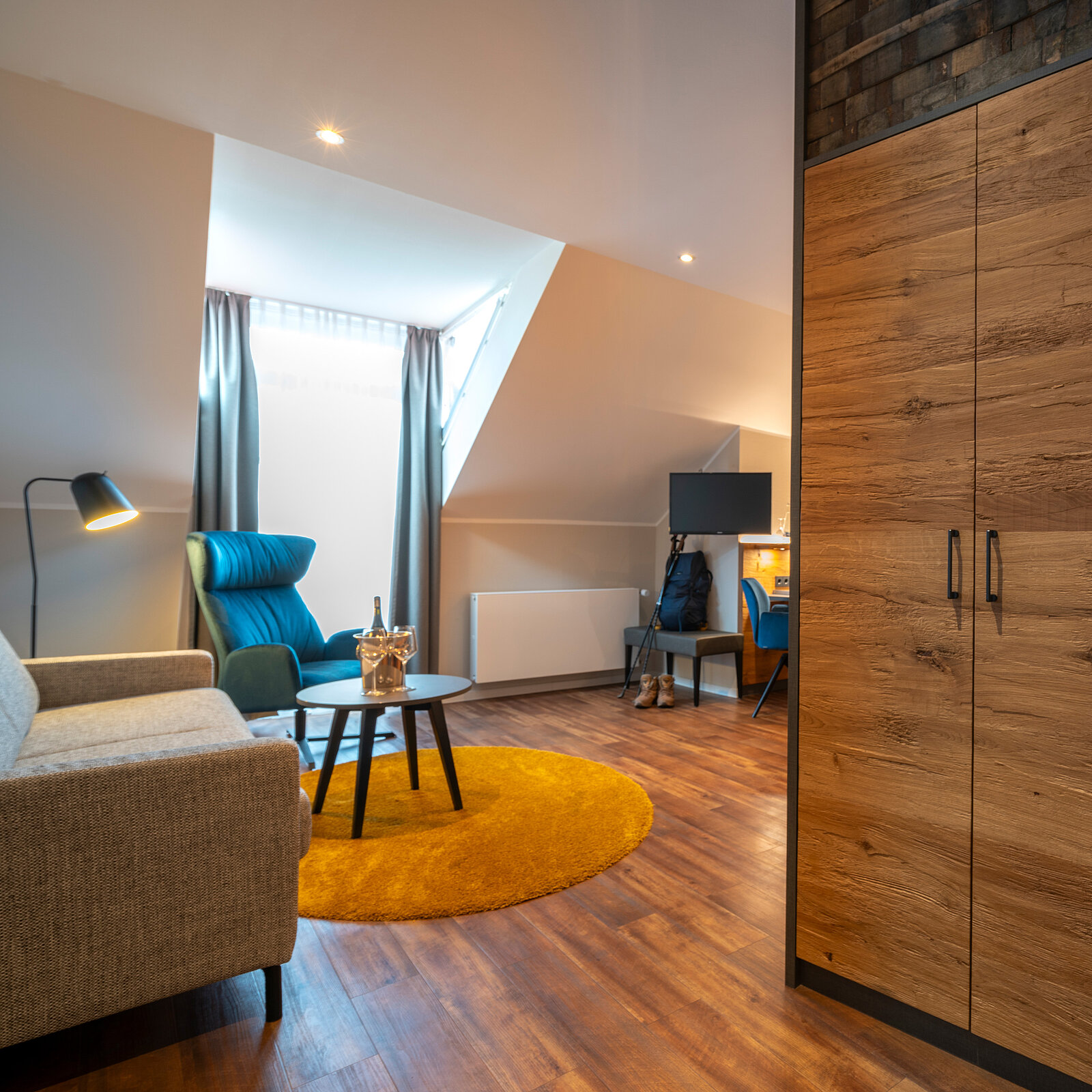 Ein helles Zimmer mit Holzfußboden. Links steht ein graues Sofa neben einem kleinen Tisch. In der Ecke befindet sich ein blauer Sessel mit Kissen, davor ein runder Teppich. Ein Schreibtisch mit Stuhl ist im Hintergrund sichtbar, vor einem Fenster mit Vorhängen.