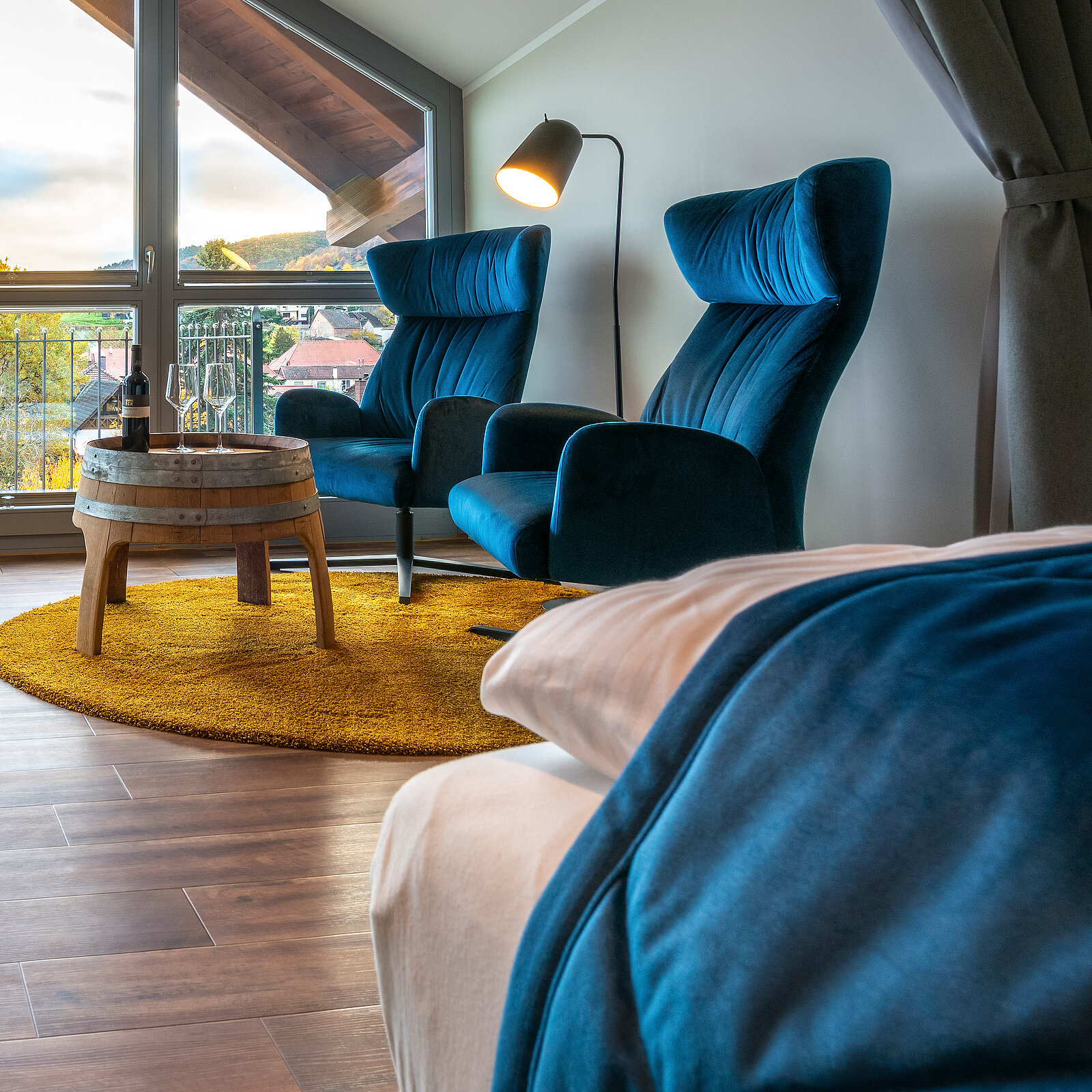 Ein gemütlicher Raum mit zwei blauen Sesseln, einem runden Tisch aus einem Fass und einem gelben Teppich. Große Fenster bieten einen Ausblick auf die Landschaft. Eine Stehlampe sorgt für Beleuchtung. Der Raum strahlt eine einladende und entspannte Atmosphäre aus.