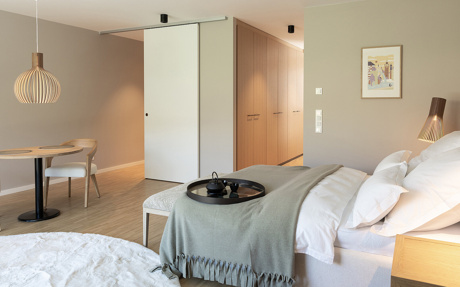 Modernes Schlafzimmer in neutralen Tönen mit Doppelbett, grauer Decke, Holzlampe, rundem Tisch und Stuhl. Schiebetür öffnet sich zu einem Flur mit Kleiderschrank. Minimalistische Einrichtung mit einem Bild an der Wand.