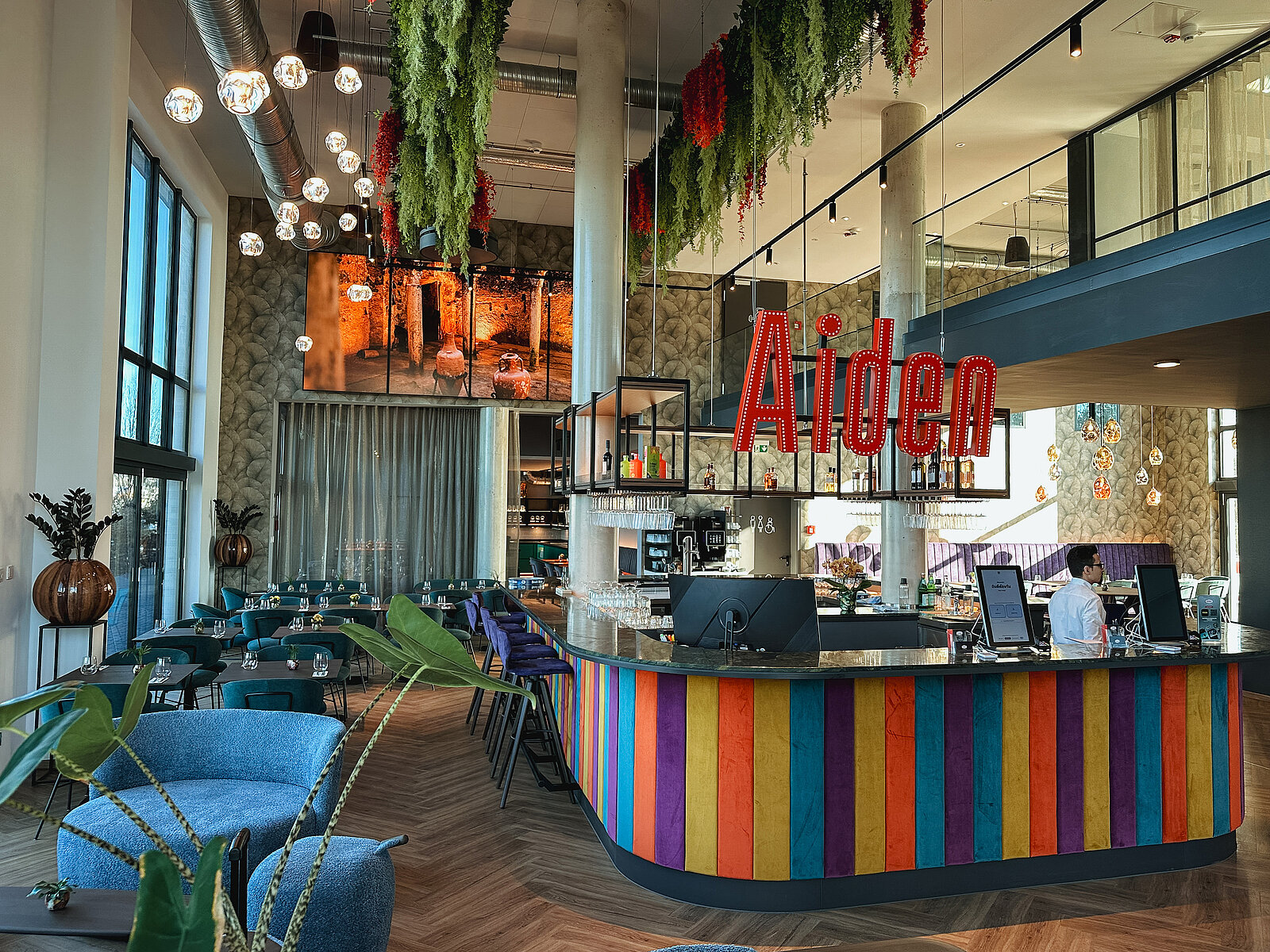 Modernes Café oder Restaurant mit bunter Theke, großen Fenstern und viel Tageslicht. Pflanzen hängen von der Decke, und Menschen arbeiten an Computern hinter der Theke. Ambiente wirkt stilvoll und einladend, mit einer auffälligen roten Neonschrift "Aiden" über der Theke.