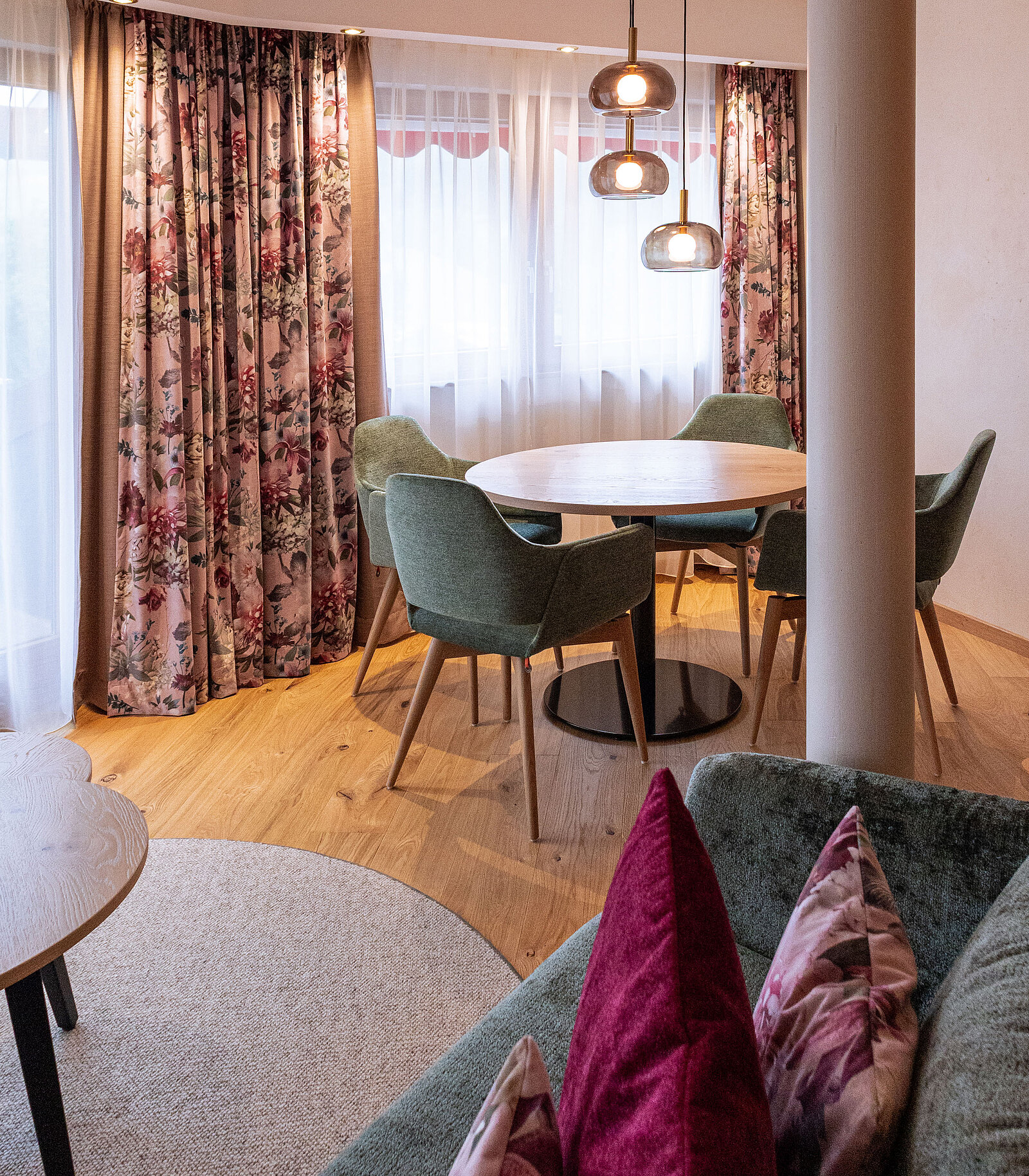 Gemütlicher Essbereich mit einem runden Holztisch und vier grünen Stühlen auf einem hellen Holzboden. Große Fenster mit geblümten Vorhängen sorgen für viel Licht. Moderne, hängende Lampen verleihen dem Raum eine warme Atmosphäre.