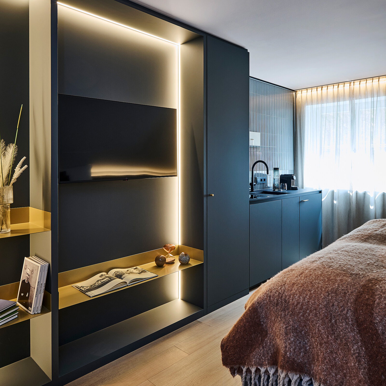 Ein modernes Hotelzimmer mit einem großen Flachbildfernseher in einem beleuchteten Regal, daneben zwei Schränke. Eine Küchenzeile mit Kaffeemaschine ist sichtbar. Vor einem großen Fenster mit Vorhängen steht ein gemütliches Bett mit einer braunen Decke.