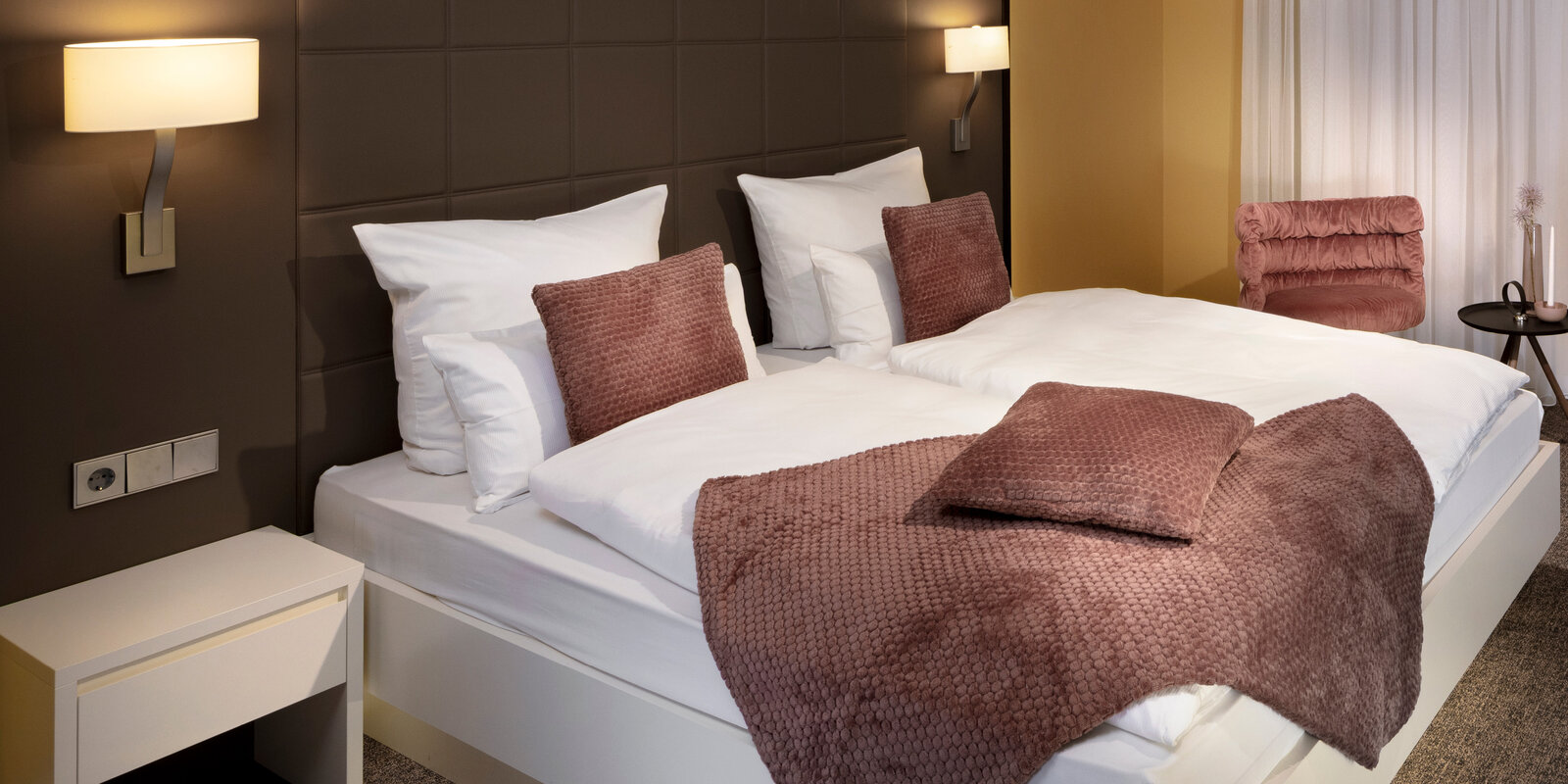 Modern hotel room with two neatly made beds, brown accent pillows and throws, wall-mounted lights, and a small side table with a chair.