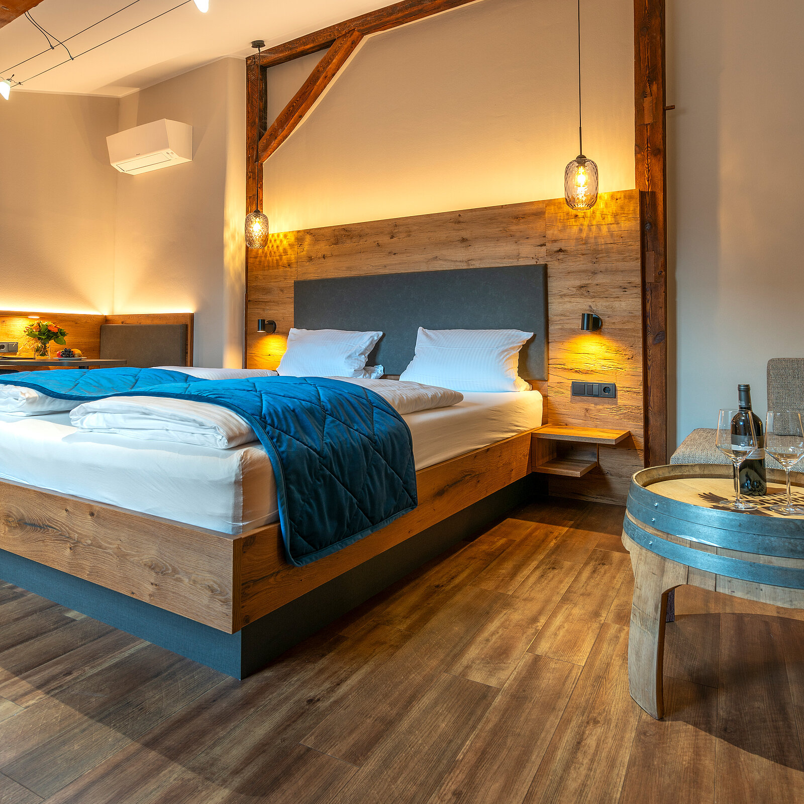 Ein modern eingerichtetes Schlafzimmer mit einem großen Bett aus Holz. Die Bettwäsche ist weiß, und eine blaue Decke liegt auf dem Bett. An der Wand befinden sich zwei Nachttische mit Lampen, und der Boden ist aus Holz. Eine angenehme, ruhige Atmosphäre.