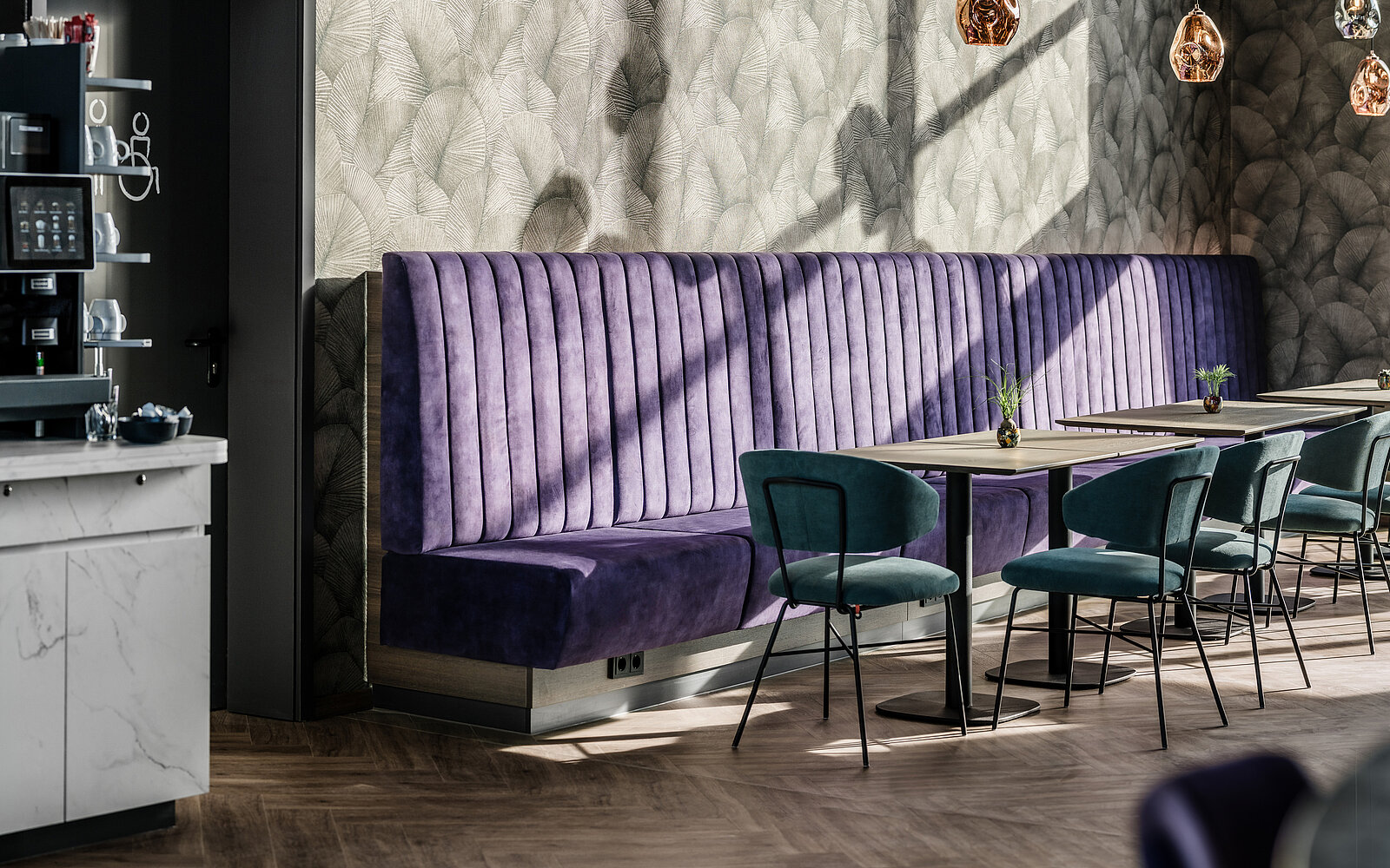 Gemütliche Sitzecke in einem Café mit lila gepolsterten Bänken und grünen Stühlen an Holztischen. Die Wand hat ein dezentes Muster, und alles ist von warmem Sonnenlicht beleuchtet.