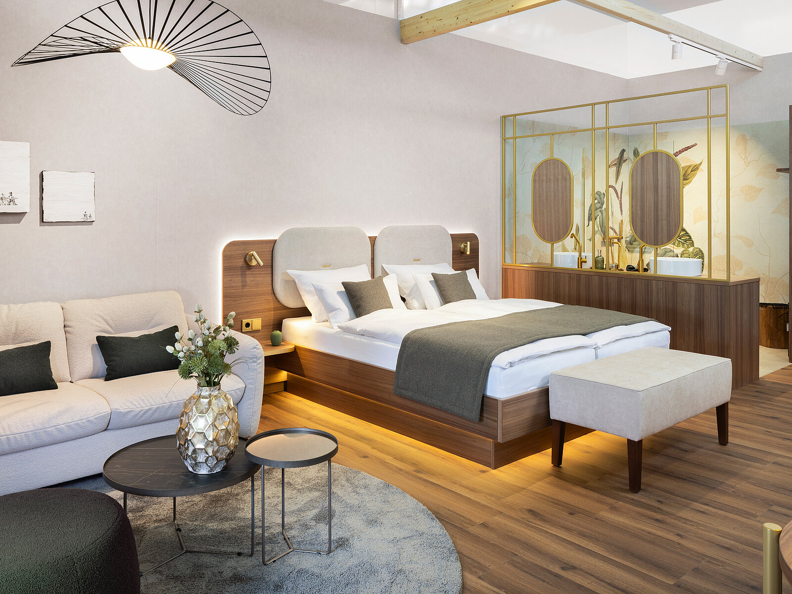 A modern hotel room featuring a wooden double bed with light bedding and a decorative headboard. A cozy sofa with cushions is nearby, accompanied by a round coffee table. The space has a stylish design with wooden flooring and elegant decor, creating a welcoming atmosphere.