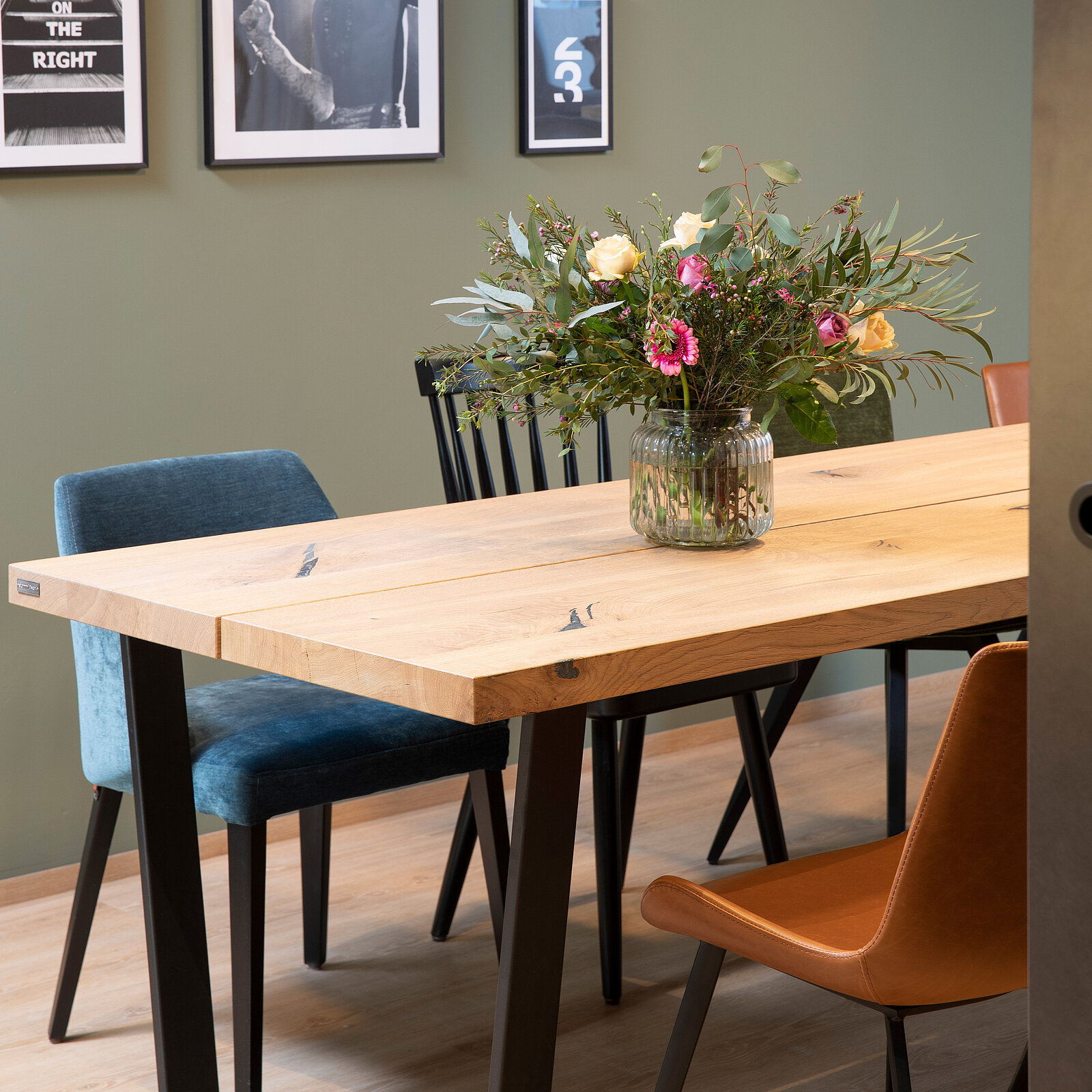 Wooden dining table with a vase of flowers, surrounded by mismatched chairs. Framed photos hang on a green wall in the background.