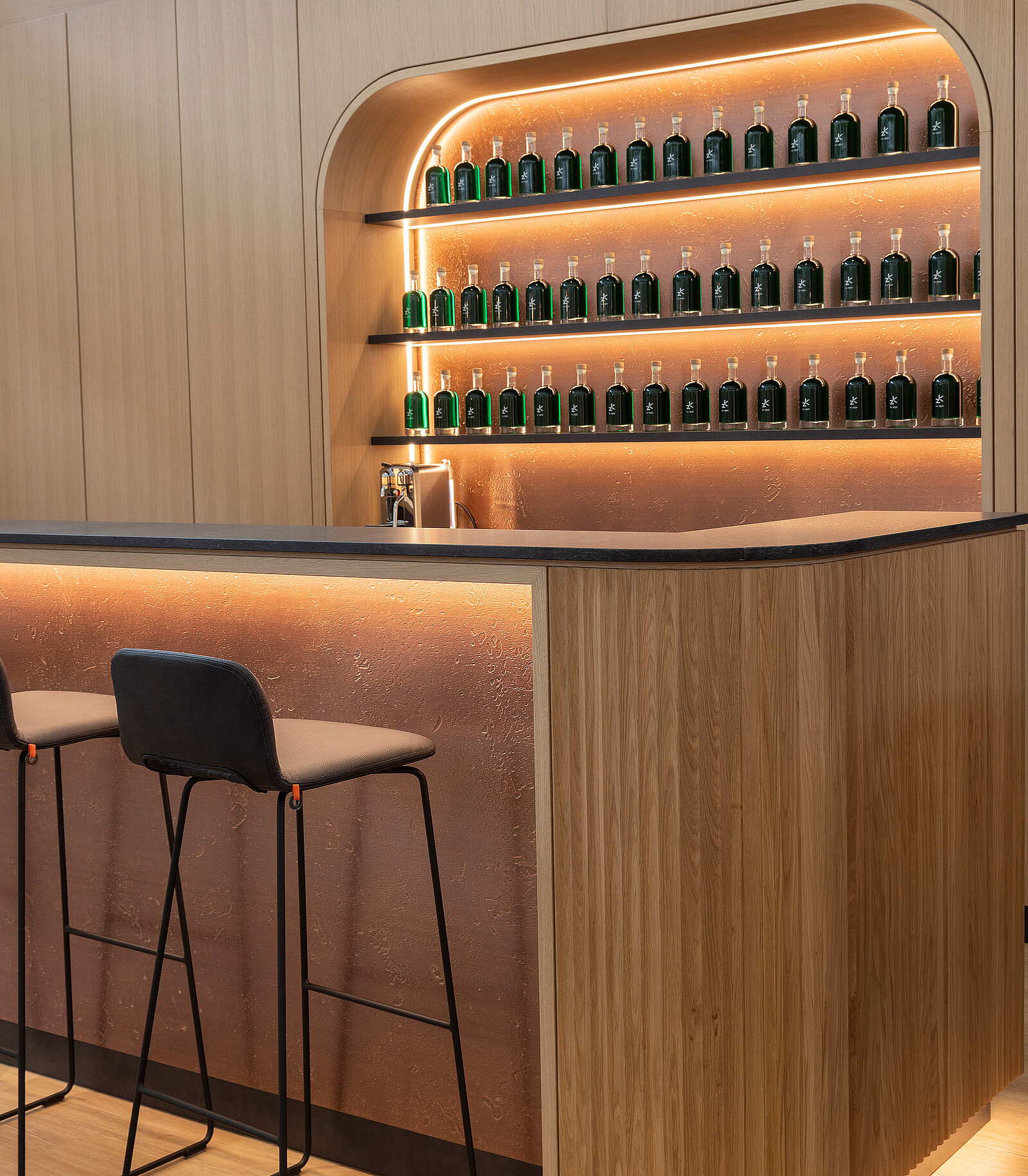 A modern bar features a curved wooden counter and two high stools. Behind the counter, shelves display numerous green glass bottles illuminated by warm lighting, creating an inviting atmosphere. The overall design emphasizes clean lines and a contemporary aesthetic.