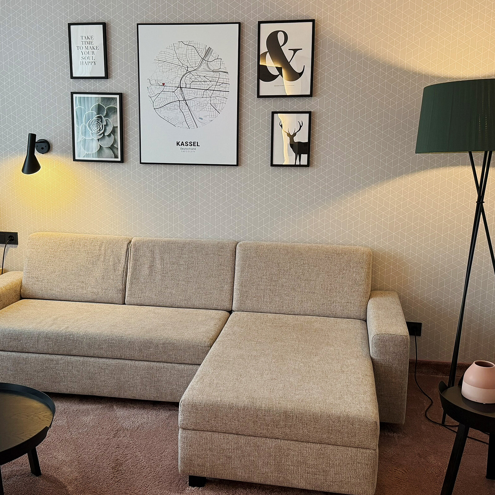 Ein gemütliches Wohnzimmer mit einem hellbeigen Sofa und einem runden, dunklen Tisch. An der Wand hängen mehrere Bilder, darunter eine Karte von Kassel. Eine Stehlampe rundet die Einrichtung ab. Der Boden ist mit einem weichen Teppich bedeckt.