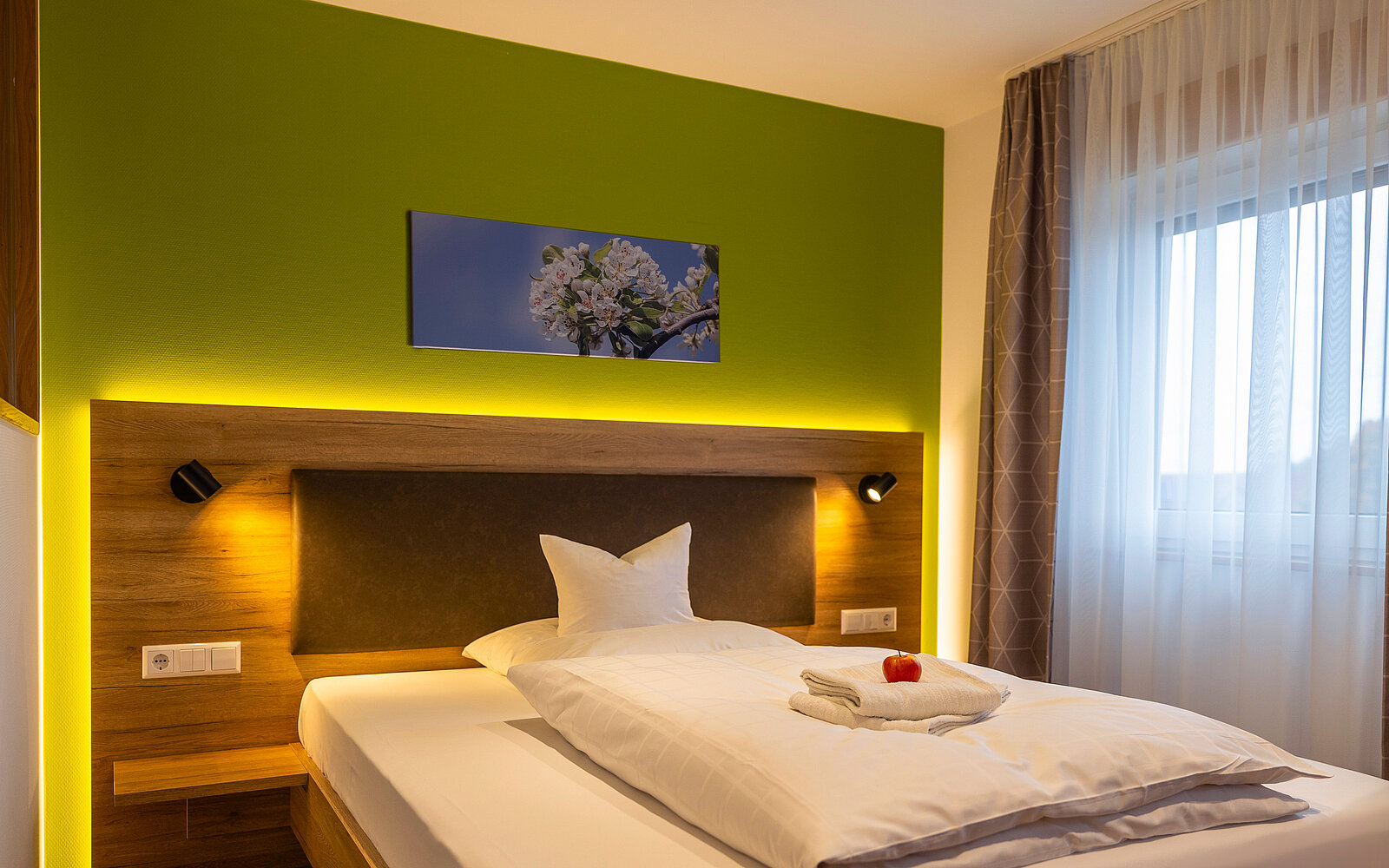 Ein modernes Schlafzimmer mit einem Einzelbett, helle Bettwäsche und ein gefaltetes Handtuch mit dekorativem Apfel darauf. Die grüne Wand mit einem Blumenbild verleiht dem Raum Farbe. Eine große Fensterfront mit halbtransparentem Vorhang lässt Tageslicht herein.