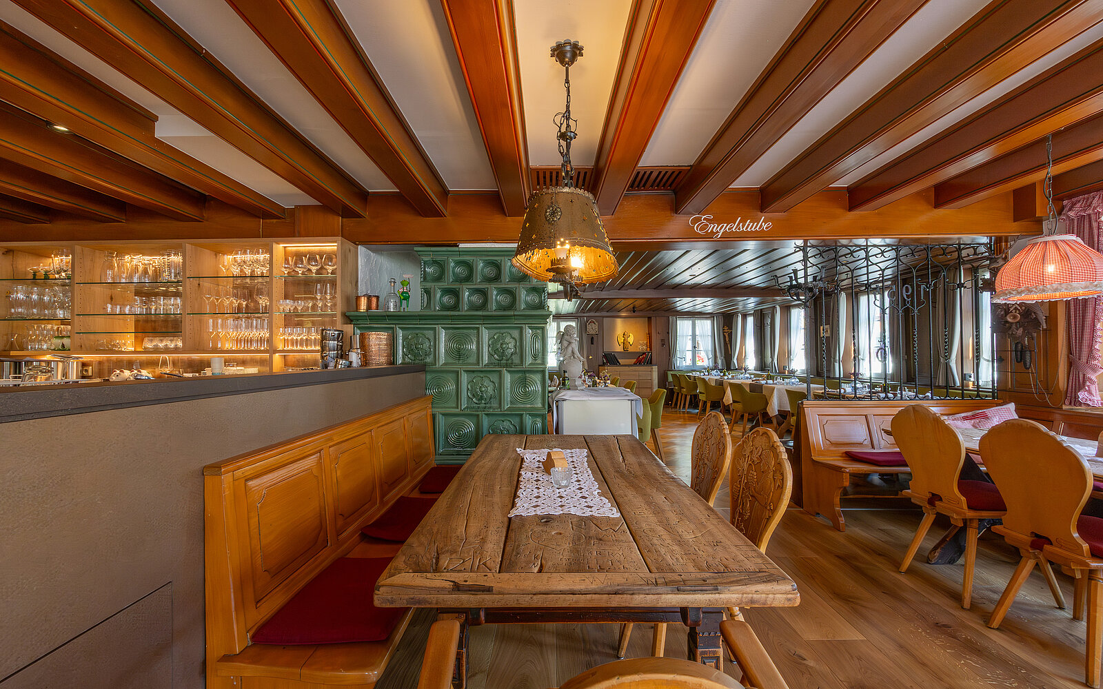 Ein gemütliches Restaurant mit Holzmöbeln, einer langen Holzbank an einem Tisch, traditionellen Stühlen und einem großen grünen Kachelofen im Hintergrund. Die Decke ist mit Holzbalken verziert.