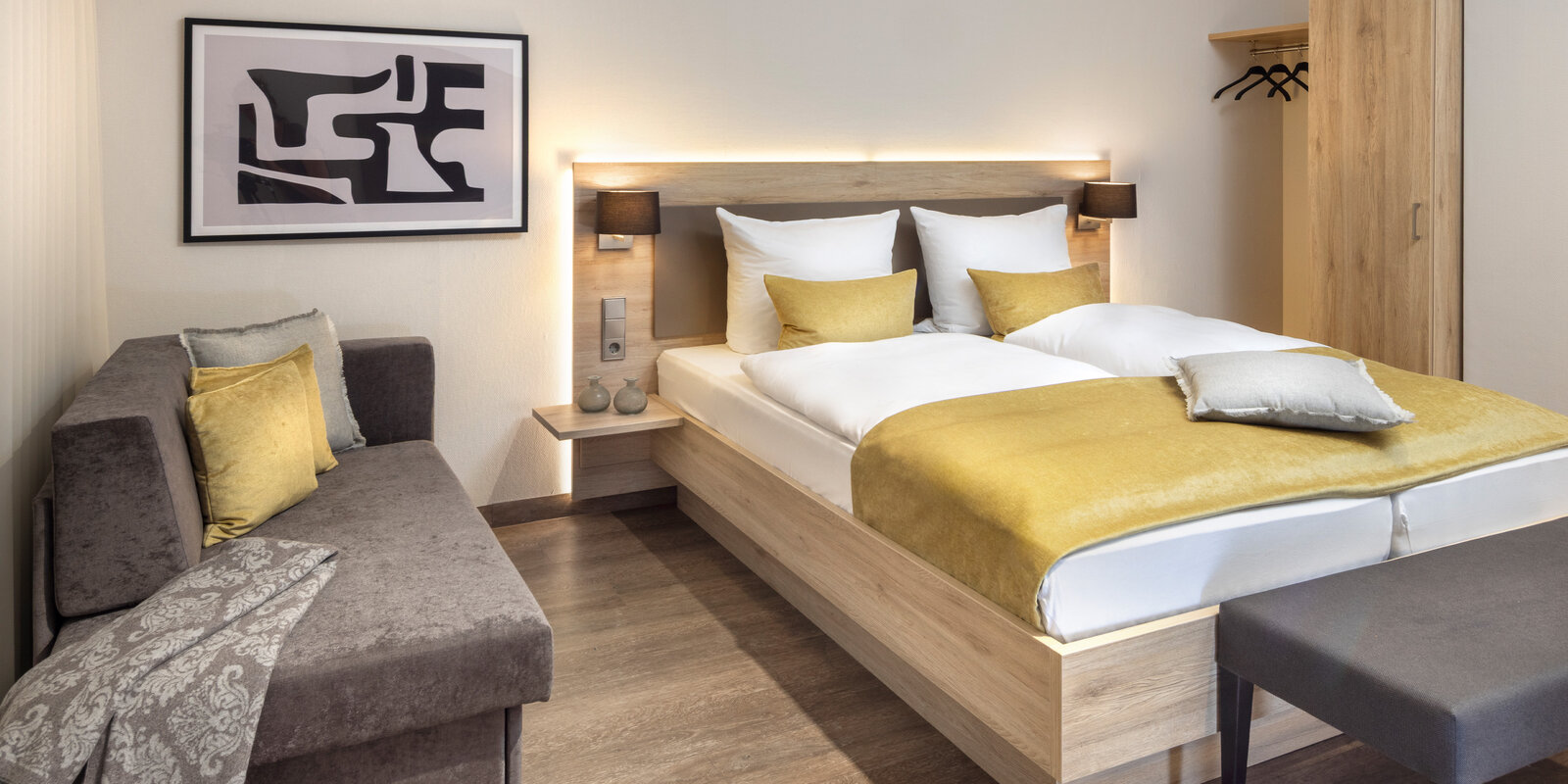 zieflekoch musterzimmer zaris, modernes Zimmer mit einem Doppelbett, gelben Akzenten, einem grauen Sofa, abstrakter Wandkunst und einem Holzschrank, die warme Beleuchtung schafft eine gemütliche Atmosphäre