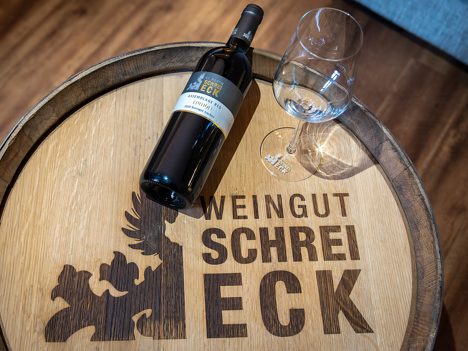Auf einem Holztisch liegt eine Flasche Wein des Weinguts Schreieck, daneben steht ein Weinglas. Der Tisch ist mit dem Logo des Weinguts graviert.