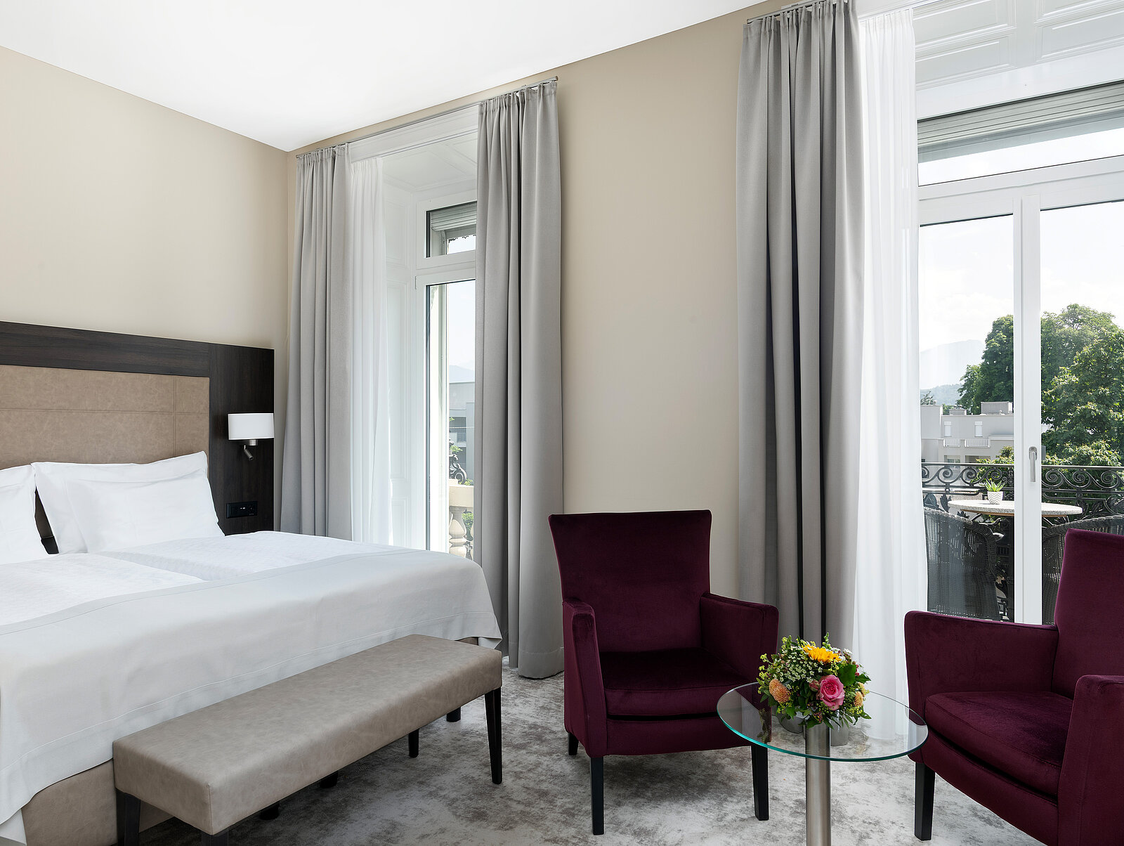 Ein elegantes Hotelzimmer mit einem großen Bett, zwei lila Sesseln, einem kleinen Glastisch mit Blumenarrangement und bodentiefen Fenstern mit grauen Vorhängen. Die Einrichtung wirkt modern und einladend.
