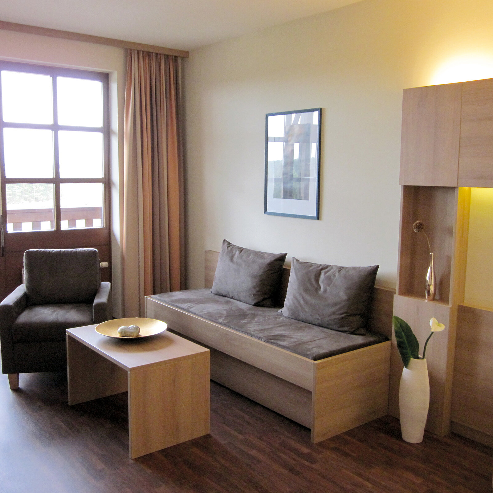 Modern living room with a brown armchair, wooden sofa, coffee table, and a tall vase. Large window with curtains and wall art.