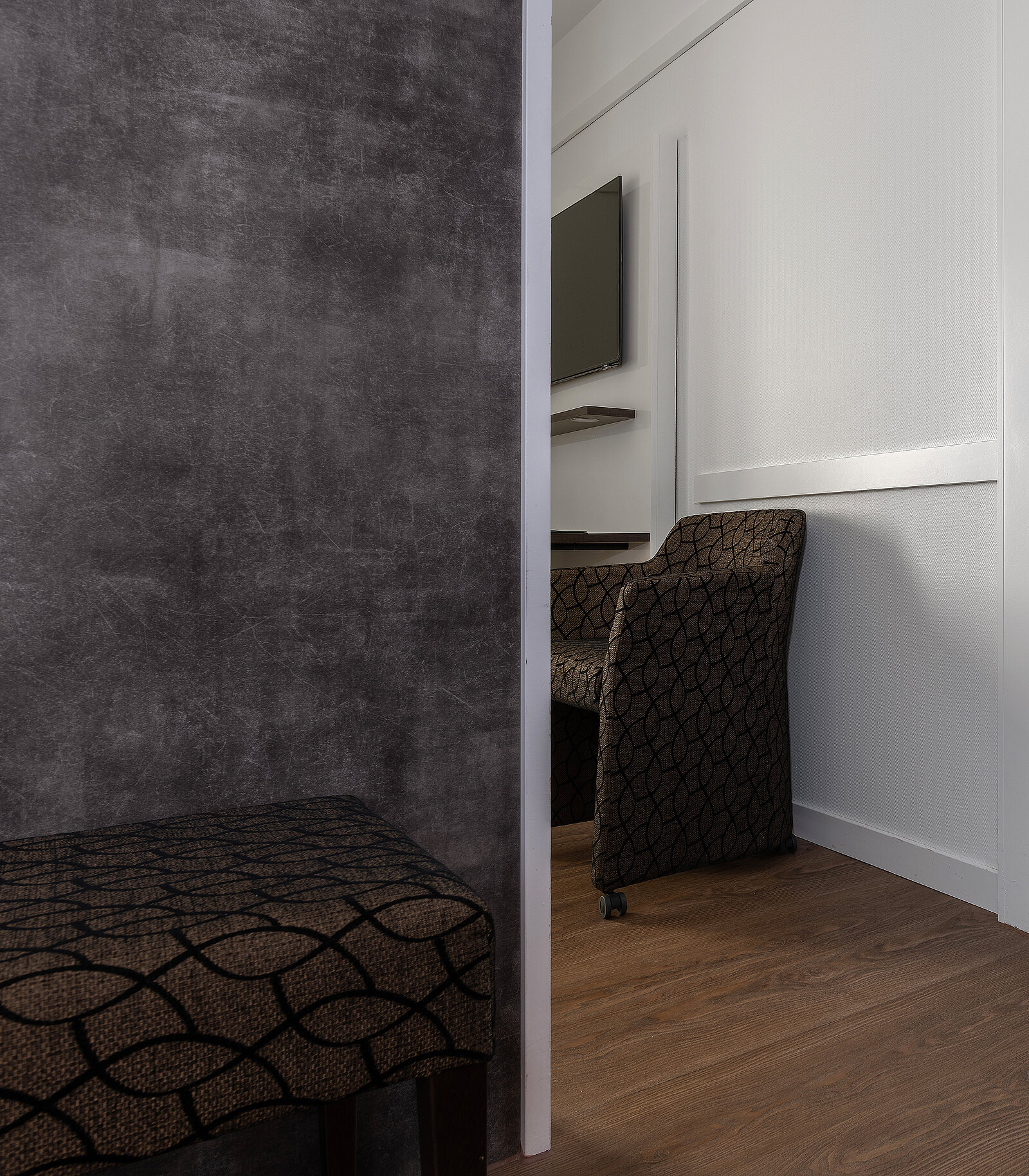 Ein modern eingerichtetes Zimmer mit einem gemusterten Sessel und Hocker, einem hölzernen Boden und einem an der Wand montierten Flachbildfernseher. Die Wand ist teilweise sichtbar und teilt das Bild in zwei Bereiche.