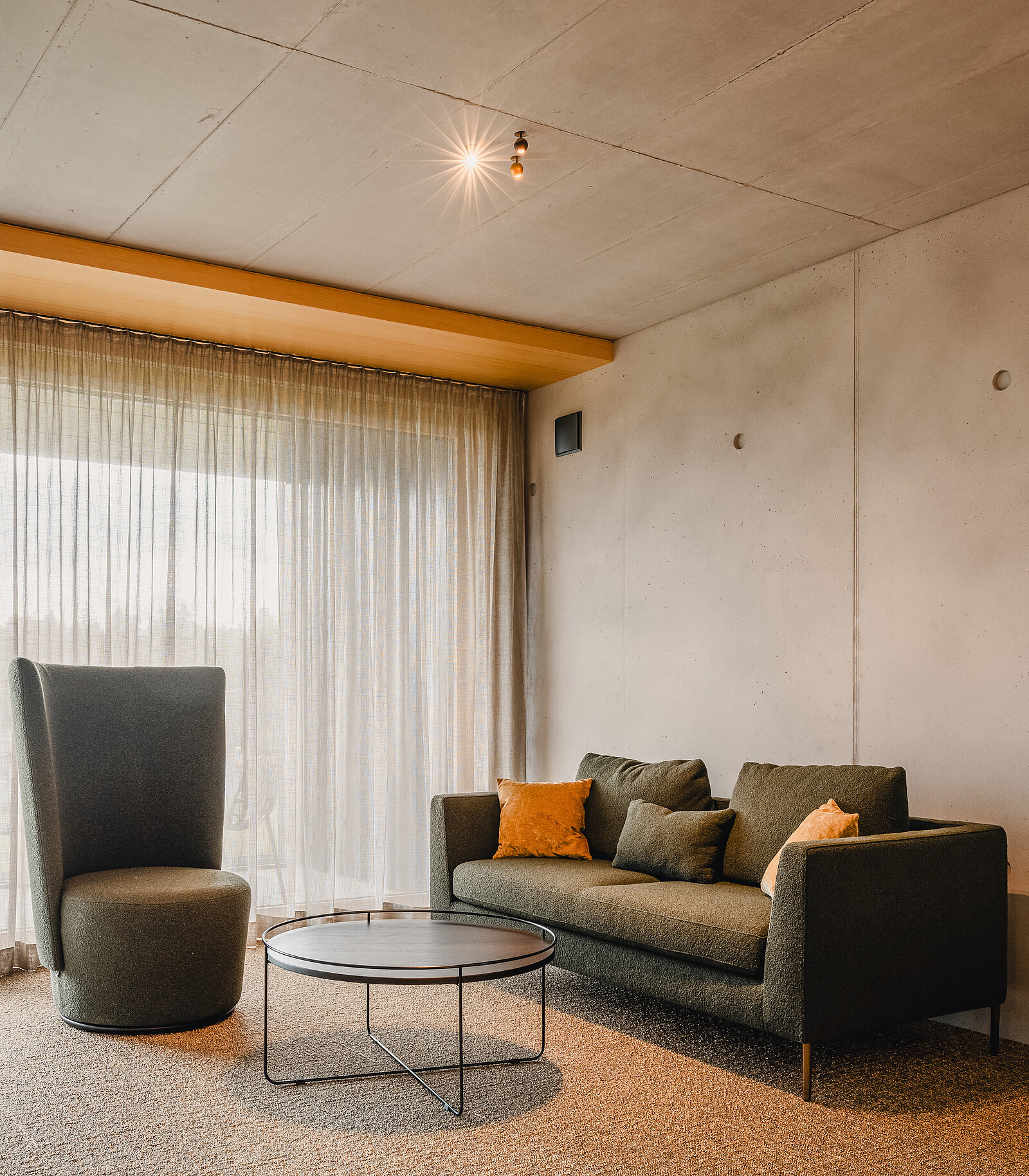 Modernes Wohnzimmer mit einem dunklen Sofa, zwei orangefarbenen Kissen und einem eleganten Sessel. Ein runder Couchtisch steht in der Mitte des Raumes, der von natürlichen Licht durch eine große Fensterfront mit Vorhängen erhellt wird. Minimalistisches Design in warmen Farbtönen.