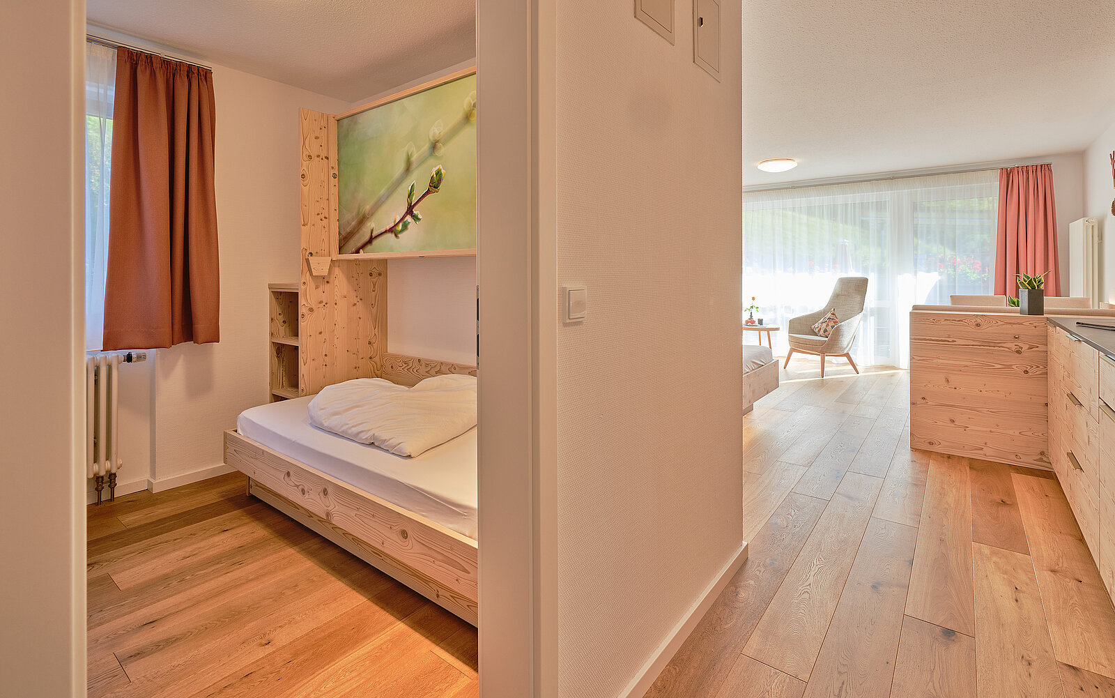 Ein modernes Apartment mit Holzmöbeln: Links ein Einzelbett in einem kleinen Raum, rechts ein heller Wohnbereich mit Sessel und großem Fenster. Holzfußboden zieht sich durch beide Bereiche.