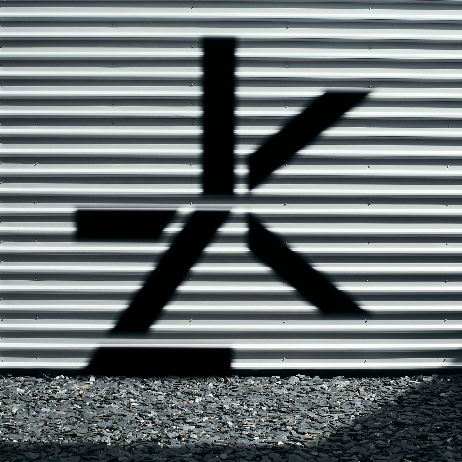 Der Schatten eines deutlichen grafischen Symbols, das einem stilisierten Buchstaben ZK ähnelt, wird auf eine strukturierte Wellwand geworfen. Der Schatten weist eckige Linien und Formen auf, die einen auffälligen visuellen Kontrast zur Oberfläche bilden. Der Hintergrund besteht aus dunklem Kies.