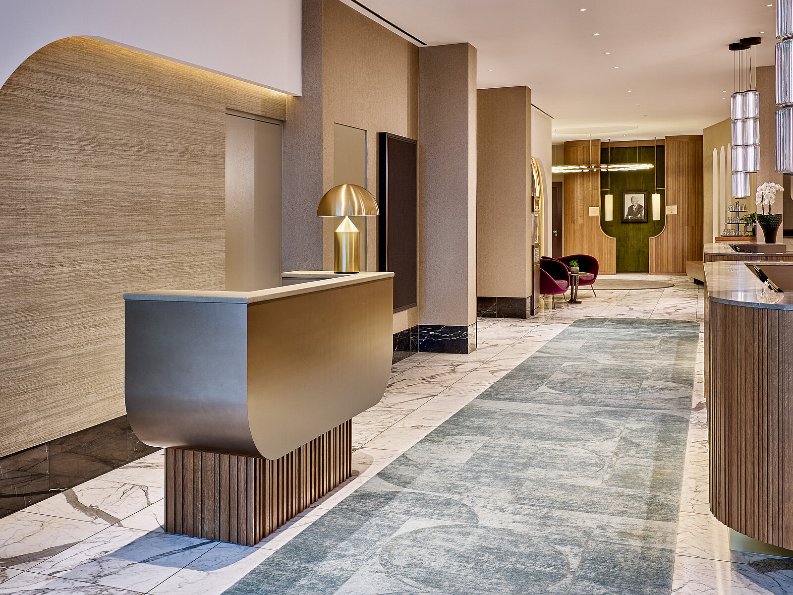 Elegante Hotel-Lobby mit modernem Empfangstresen, metallischer Lampe und einladendem Ambiente. Der Boden ist mit einem blauen Teppich ausgelegt, es gibt moderne Sitzgelegenheiten im Hintergrund. Die Beleuchtung ist warm und sorgt für eine gemütliche Atmosphäre.