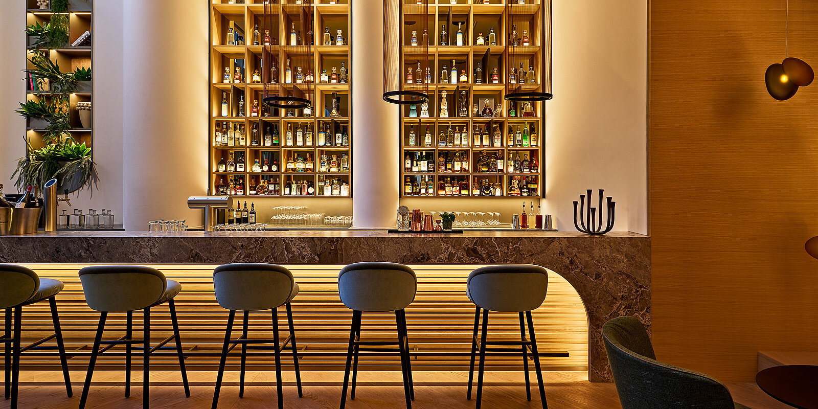 Elegante Bar mit fünf hohen Stühlen vor einer langen Theke. Im Hintergrund sind beleuchtete Regale mit verschiedenen Flaschen zu sehen. Die Atmosphäre wirkt warm und einladend.