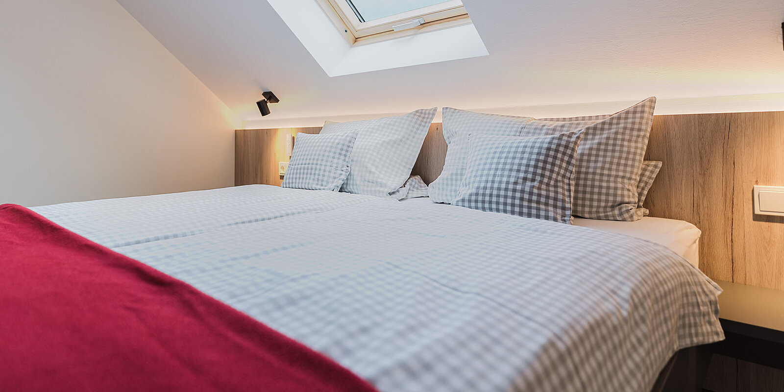 Ein gemütliches Schlafzimmer mit einem Doppelbett, das weiß-grau karierte Bettwäsche und ein rotes Plaid hat. Über dem Bett befindet sich ein Dachfenster, durch das Tageslicht einfällt.