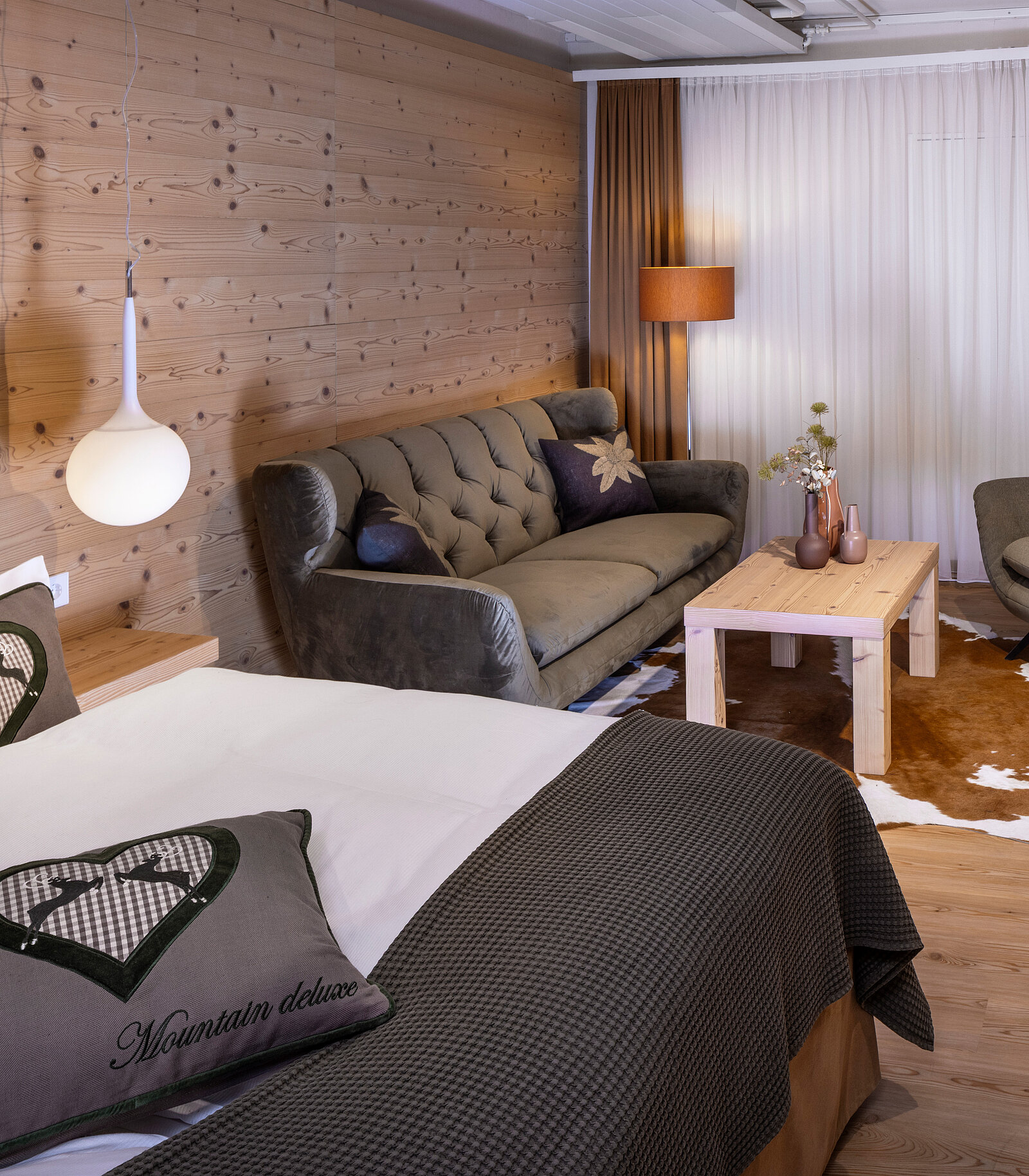 Das Bild zeigt ein stilvolles Zimmer mit einer gemütlichen Doppelbett und einer Sitzecke. Die Wände sind aus Holz, und es gibt ein Sofa sowie einen Tisch. Eine Stehlampe und eine Hängeleuchte sorgen für angenehmes Licht. Dekorative Elemente verleihen dem Raum eine einladende Atmosphäre.