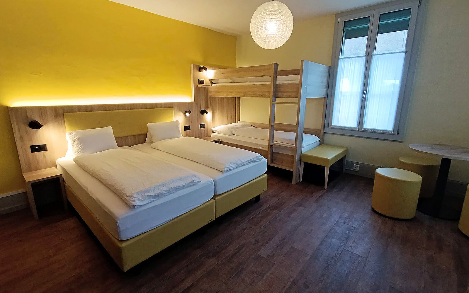 Das Bild zeigt ein helles, modernes Hotelzimmer mit zwei Einzelbetten und einem Etagenbett. Die Wände sind gelb gestrichen und die Möbel sind aus hellem Holz. Es gibt einen runden Tisch und zwei Sitzgelegenheiten in der Nähe eines Fensters.