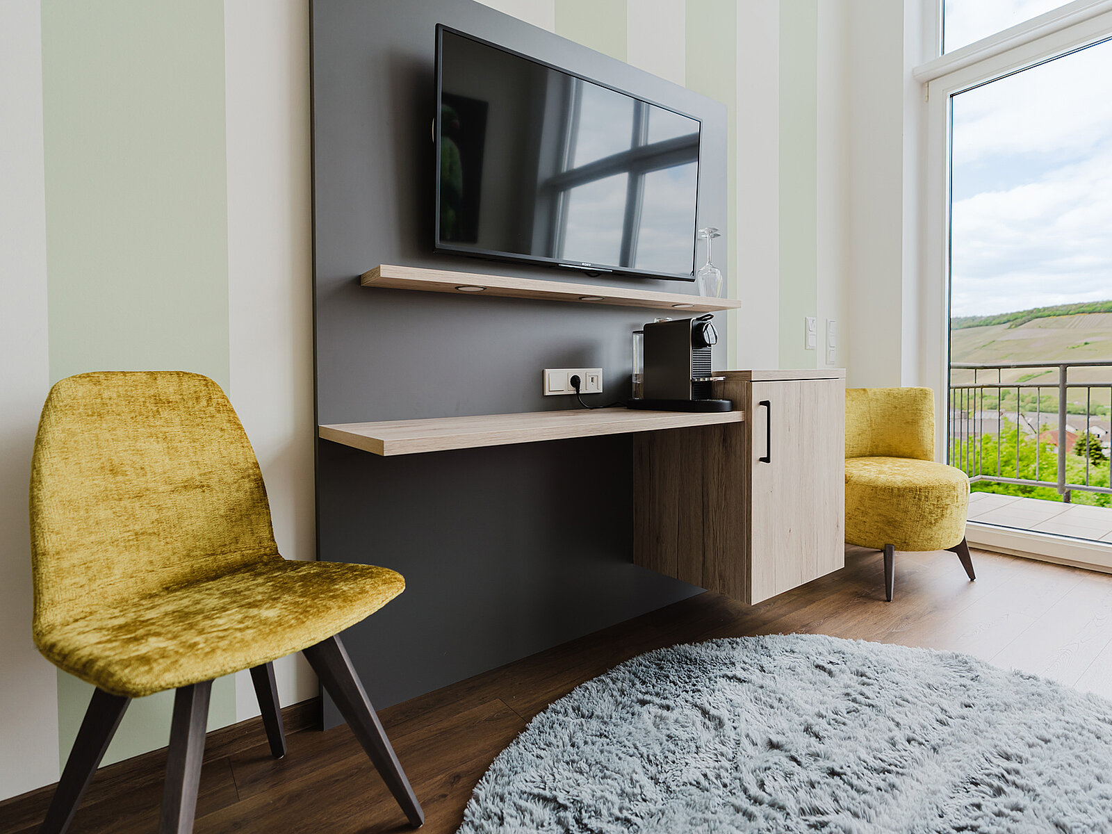 Ein modernes Zimmer mit einem Flachbildfernseher an der Wand, einem Regal darunter, zwei gelben Stühlen und einem grauen Teppich. Ein Fenster bietet Blick auf grüne Hügel.