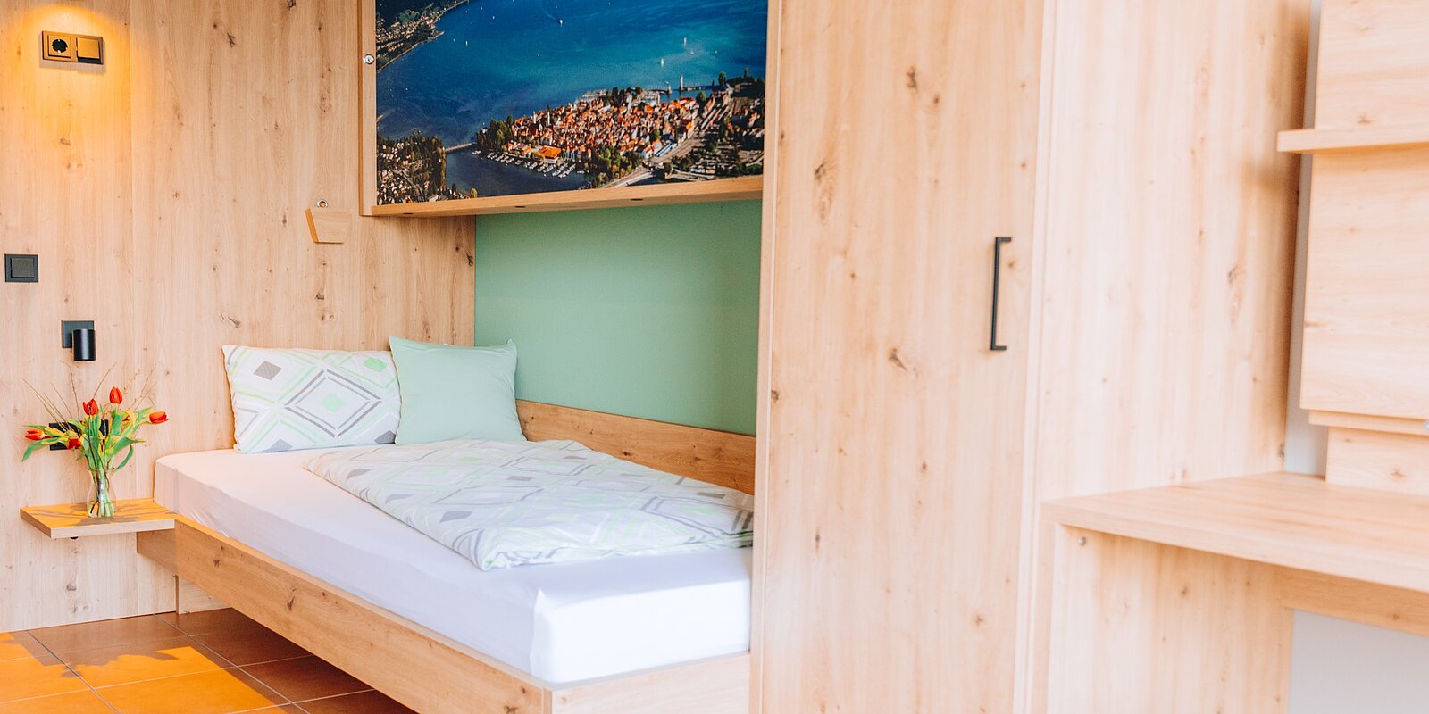 Ein modernes Schlafzimmer mit einem Einzelbett, grünen Kissen und einer bunten Bettdecke. An der Wand hängt ein großes Bild einer Stadtansicht. Auf dem Nachttisch stehen frische Tulpen. Die Wände und Möbel sind aus hellem Holz.