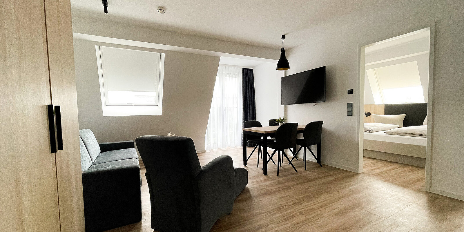 Eine moderne Wohnung mit Wohnzimmer und Schlafzimmer. Im Wohnzimmer stehen ein Sofa, ein Sessel, ein Tisch mit Stühlen und ein Fernseher an der Wand. Im angrenzenden Raum ist ein Bett sichtbar. Holzboden und große Fenster sorgen für Helligkeit.