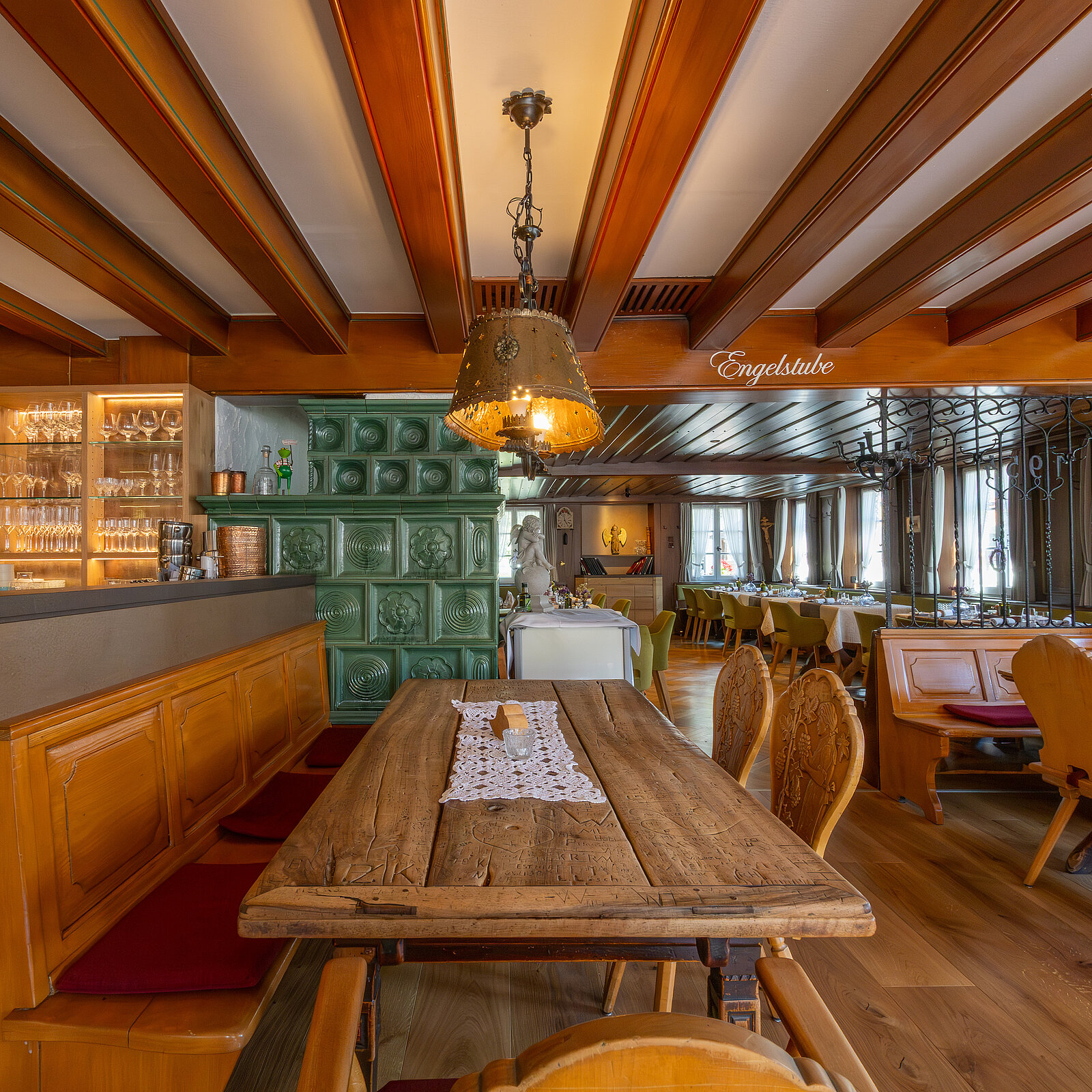 Ein gemütliches Restaurant mit Holzmöbeln, einem langen Holztisch in der Mitte und grünen Kachelofen im Hintergrund. Ein großer Kronleuchter hängt über dem Tisch. Die Atmosphäre ist warm und einladend.