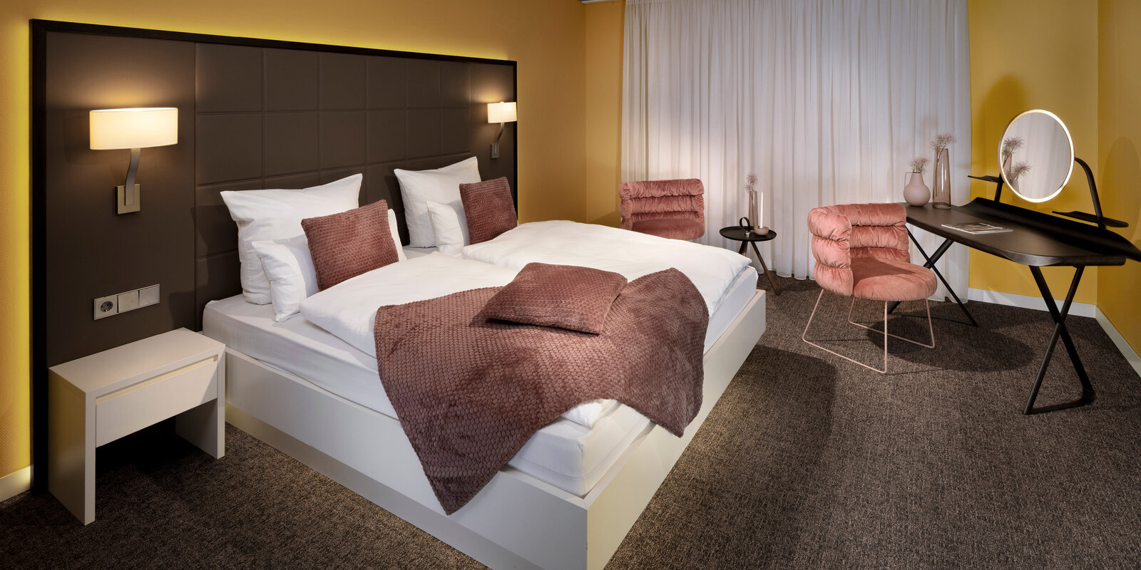Modern hotel room with a large bed, brown and pink accents, a desk with a mirror, two chairs, and soft lighting.