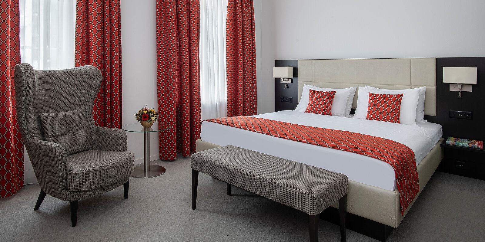 Ein modernes Hotelzimmer mit einem Doppelbett, das rote Bettwäsche und Kissen hat. Die Vorhänge passen zur Bettwäsche. Ein grauer Sessel und ein kleiner runder Tisch stehen neben dem Bett. Ein gepolsterter Hocker befindet sich am Fußende des Bettes.